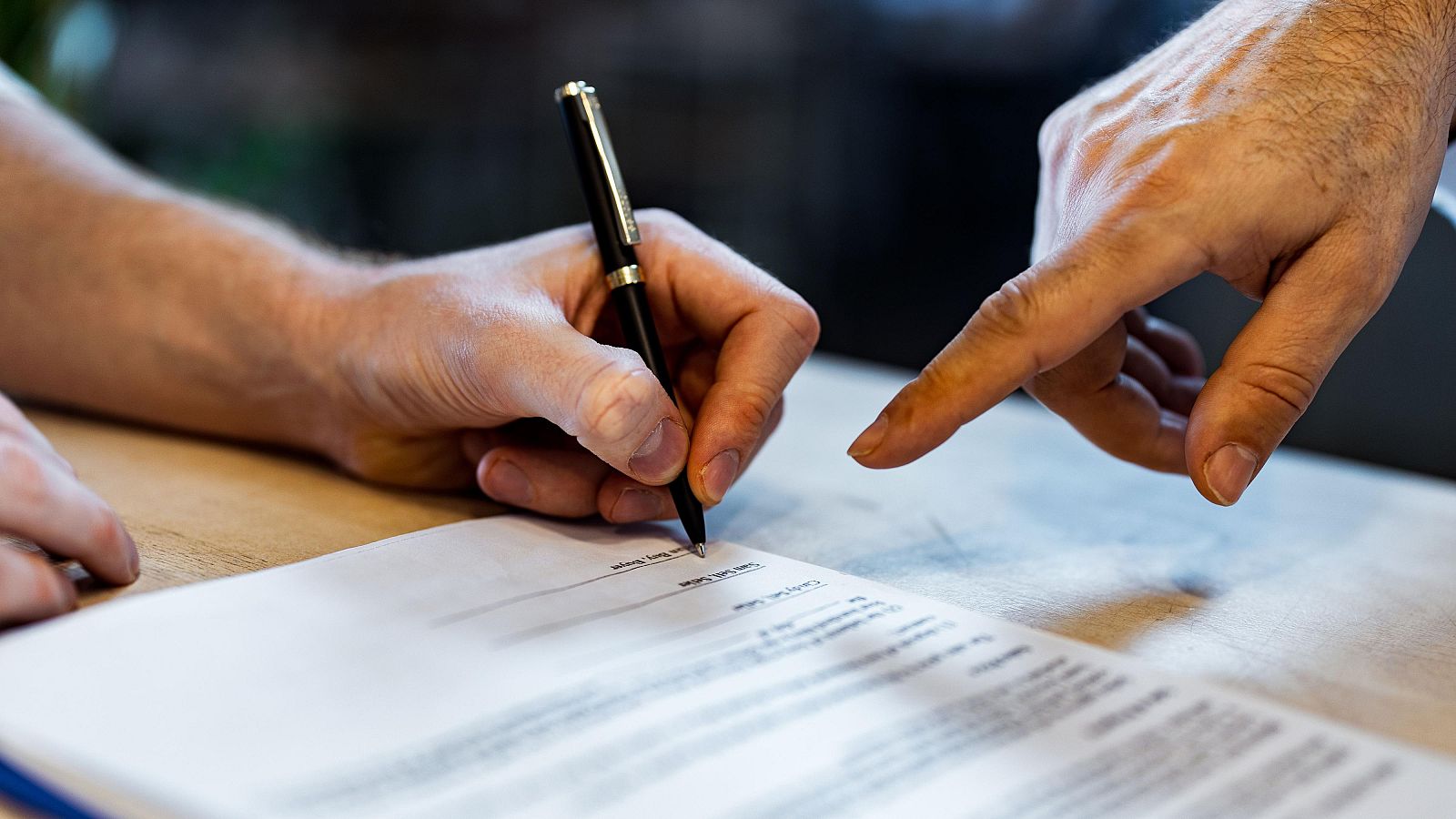 Una mano escribe con un bolígrafo sobre un documento, mientras otra mano indica un punto específico del mismo. El documento podría ser un contrato o acuerdo.