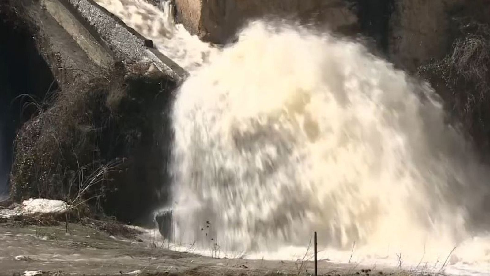 Una cascada de agua blanca cae con fuerza desde una estructura de piedra, posiblemente una presa, con vegetación escasa alrededor, creando un río en la parte inferior.