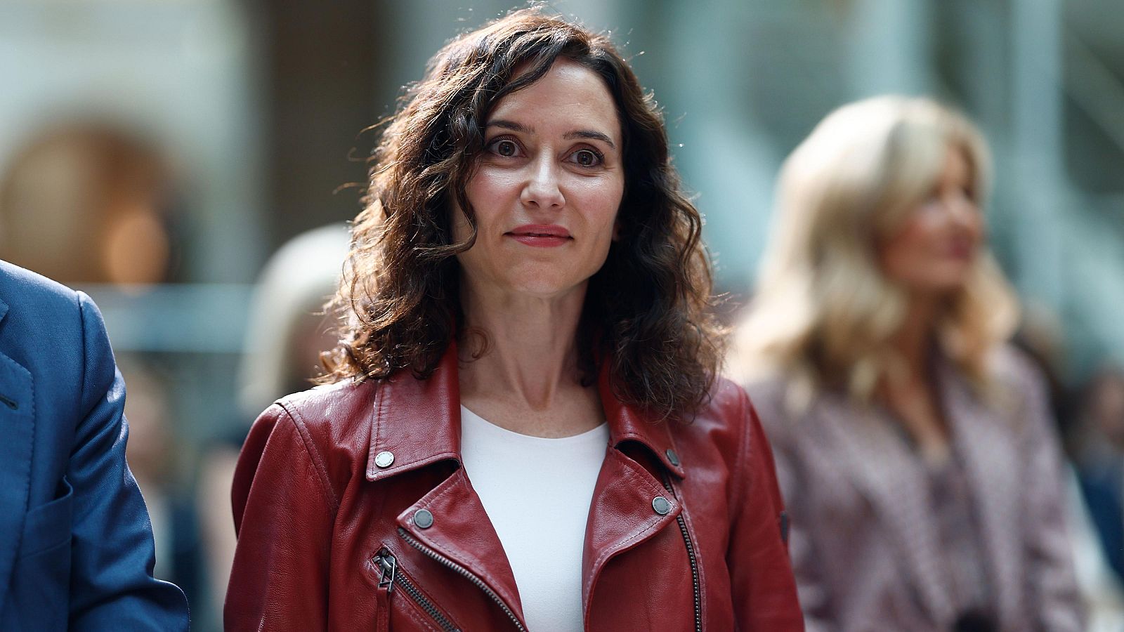 En la Asamblea, una mujer con camisa blanca y pantalones marrones se dirige a la audiencia desde un atril, gesticulando. Tras ella, se aprecia un grupo de personas sentadas, algunas prestando atención.