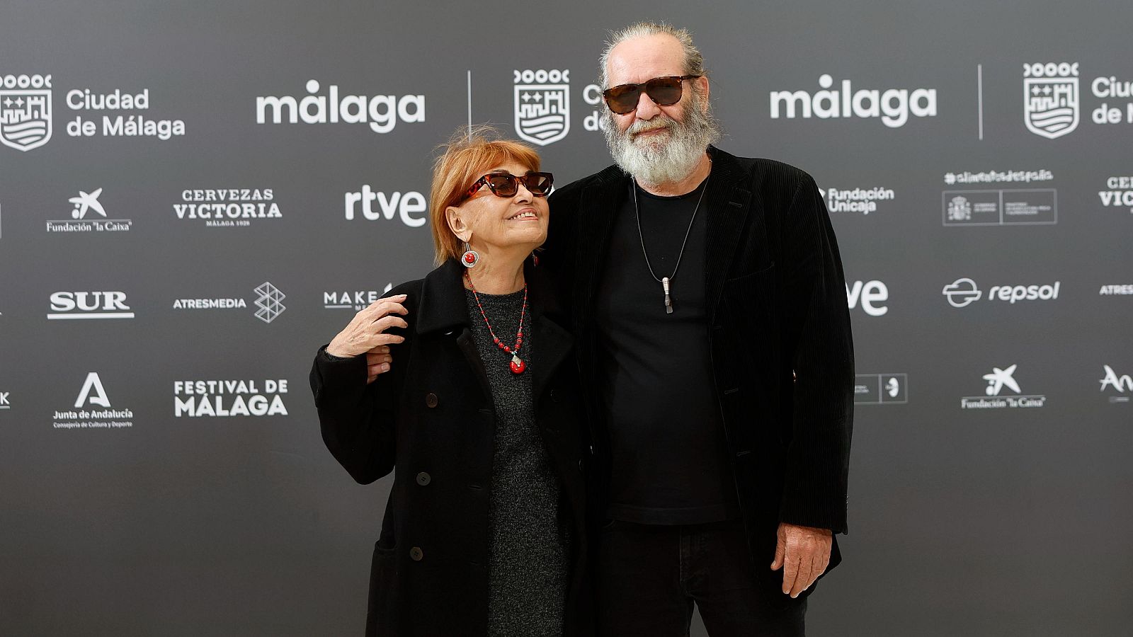 Jorge Perugorría y Mirtha Ibarra, ambos con gafas de sol, posan frente a un panel gris con logotipos. Mirtha Ibarra luce un abrigo negro y un collar rojo, mientras que Jorge Perugorría lleva una camiseta y chaqueta negras.