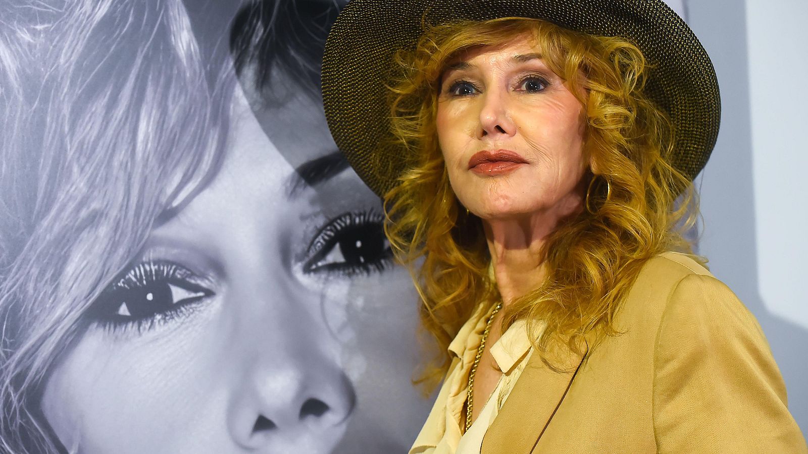 Una mujer con sombrero y traje posa frente a una imagen en blanco y negro de un rostro femenino, con un primer plano de un ojo. La mujer tiene el pelo rizado y pendientes de aro.