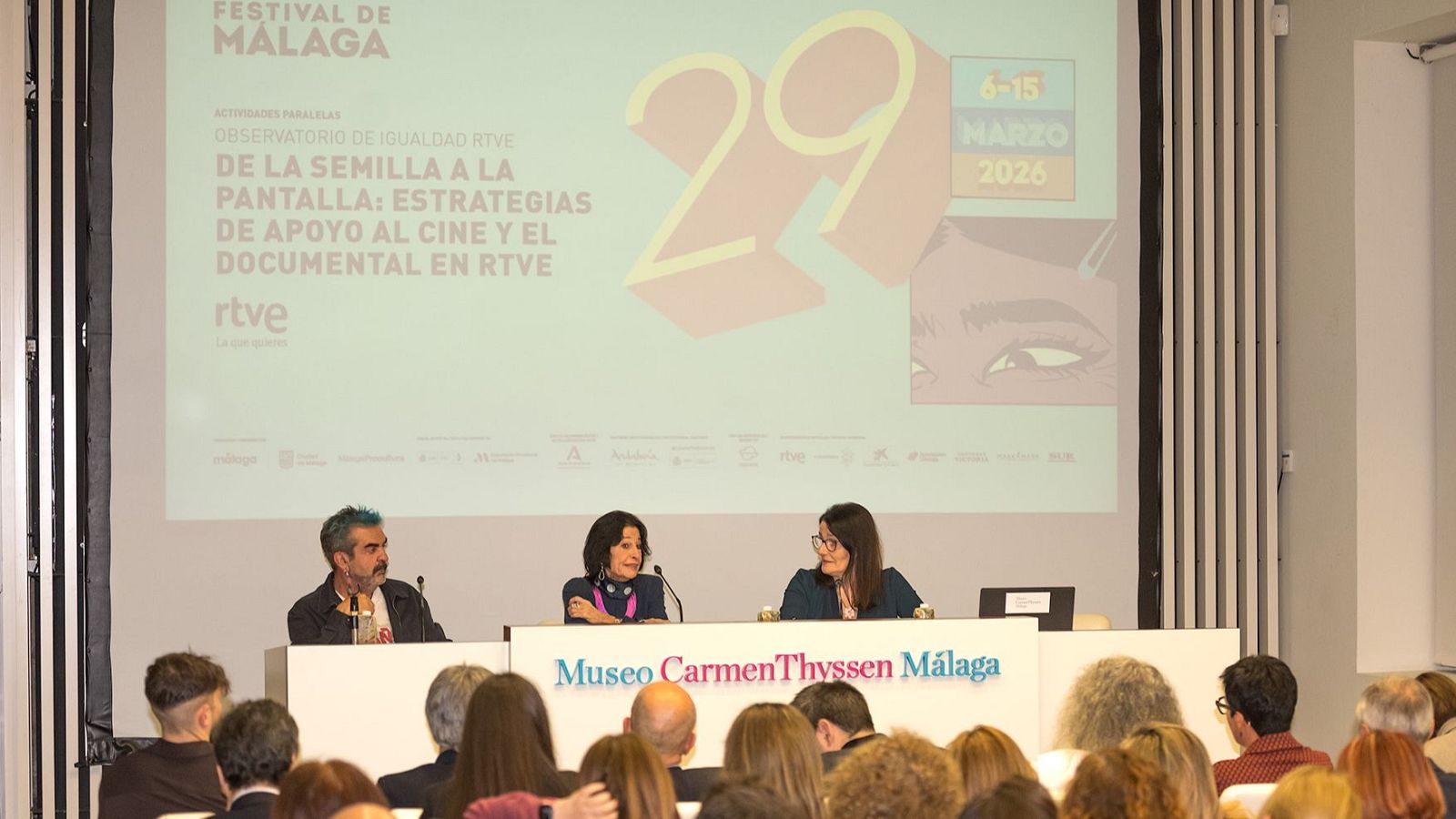 Una mesa redonda en el Museo Carmen Thyssen Málaga presenta a tres personas frente a una pantalla con logos y texto relacionados con el Festival de Málaga y RTVE, abordando estrategias de apoyo al cine y el documental.