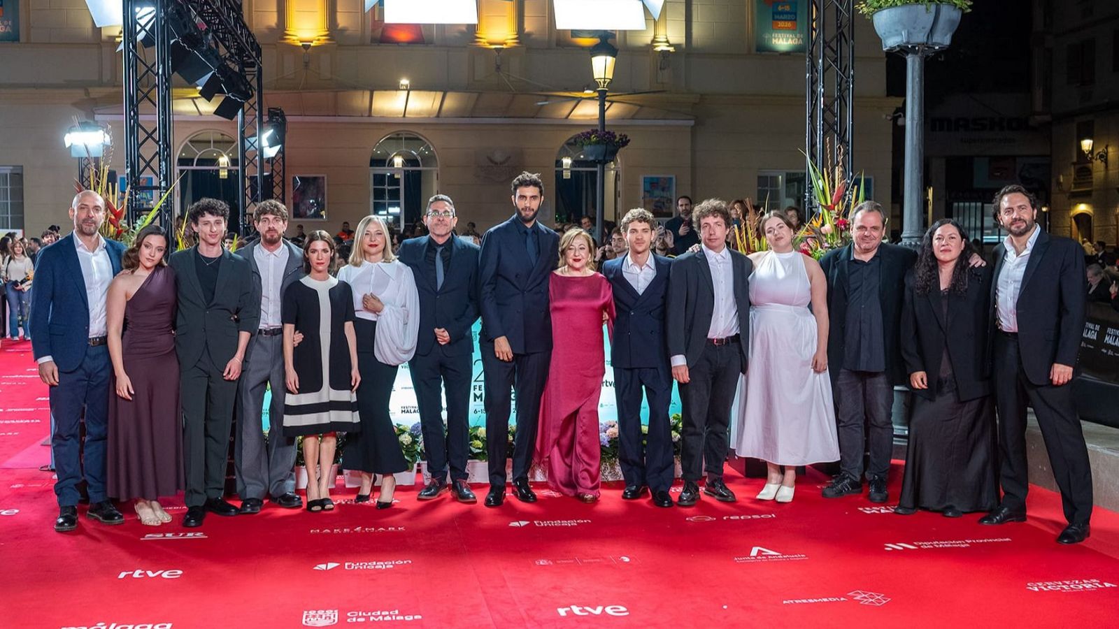 Un grupo de hombres y mujeres, elegantemente vestidos, posan en una alfombra roja frente a un edificio. Se distinguen trajes oscuros y vestidos largos, con el logotipo de un evento en el fondo.