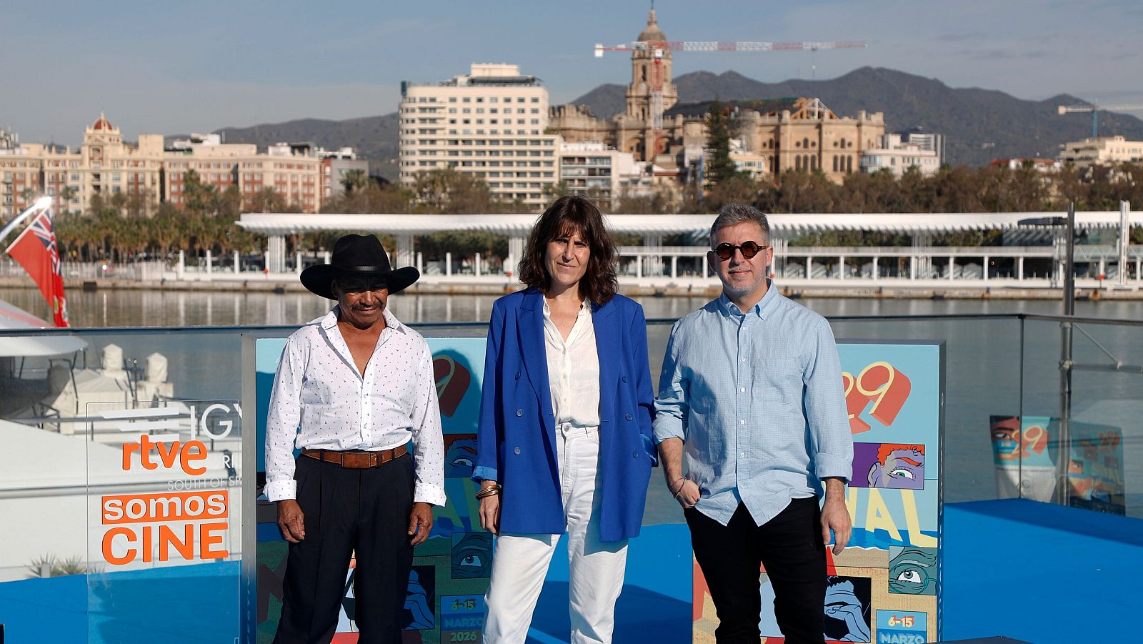 Un grupo de personas posa frente a un cartel promocional de un festival, con el skyline de una ciudad como telón de fondo. El cartel indica las fechas de celebración del evento, en marzo de 2026, y el logo de RTVE.