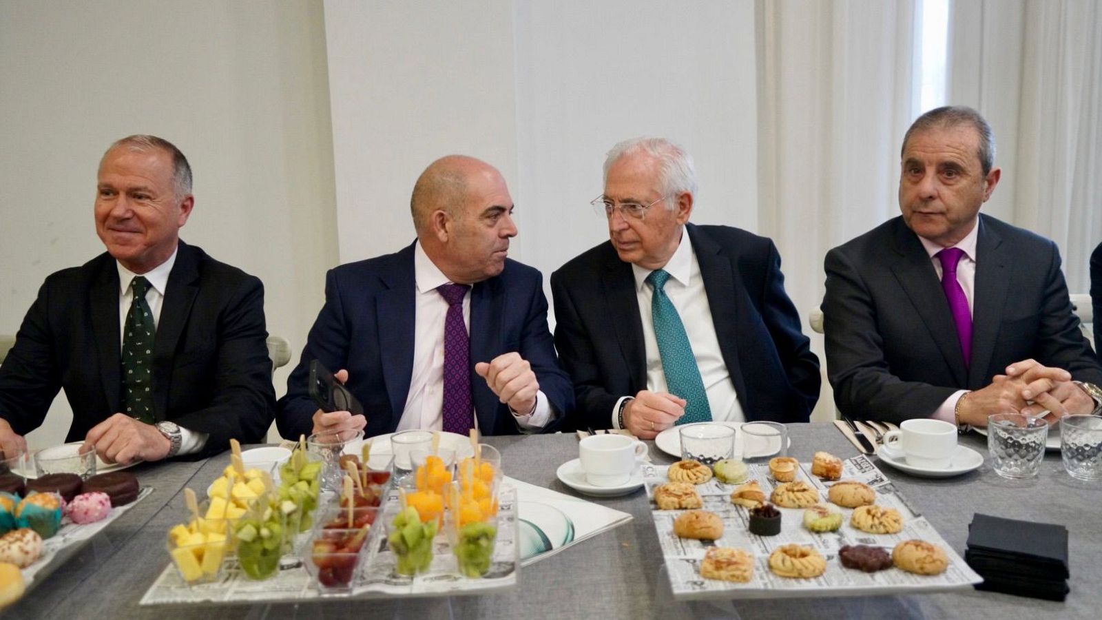 Cuatro hombres, incluyendo a Lorenzo Amor y Juan José Imbroda, se encuentran sentados alrededor de una mesa con comida y bebida. El ambiente es luminoso, con una pared blanca y una cortina clara de fondo.