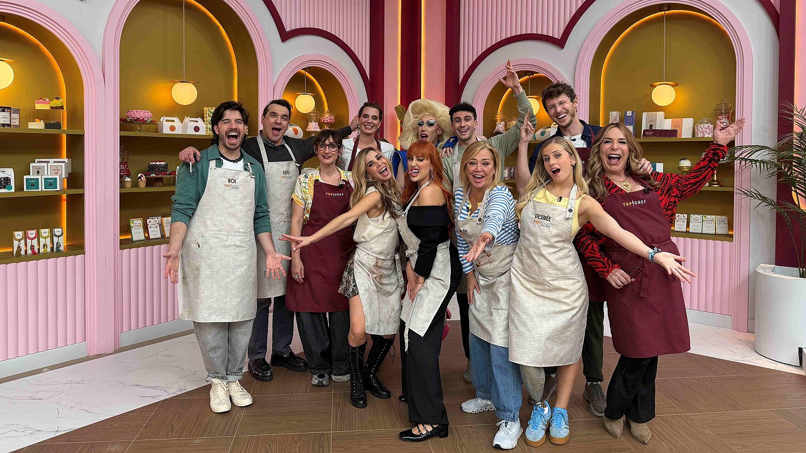 Un grupo de personas, incluyendo a una con delantal con nombre bordado y otra caracterizada, posan sonrientes en un estudio con decoración de pastelería, con un fondo rosa y dorado.