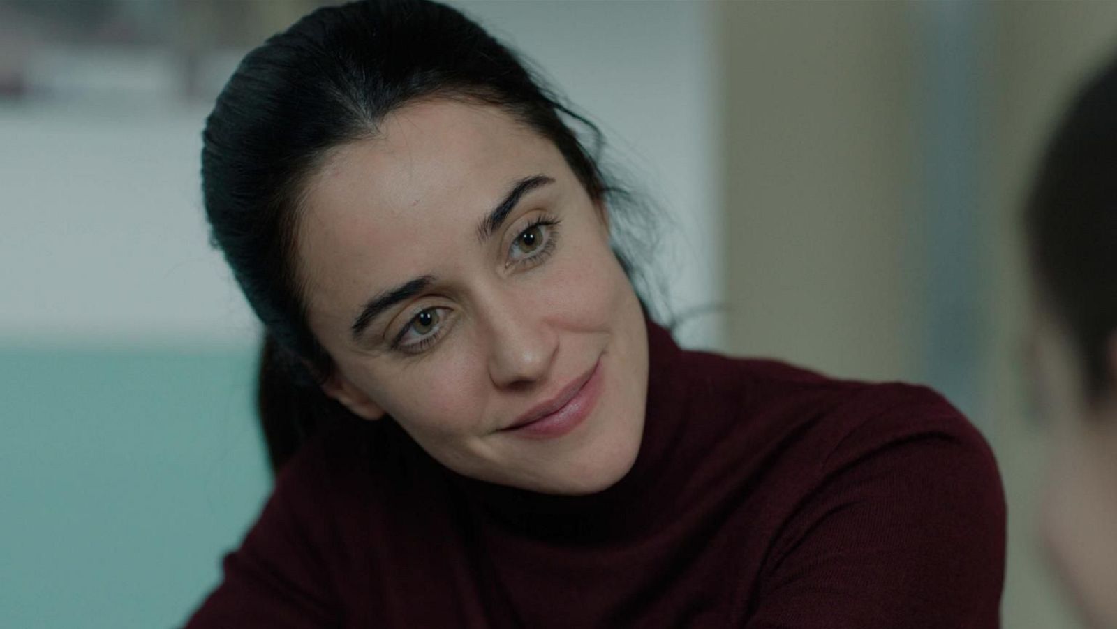 Primer plano de una mujer con cabello oscuro recogido y una sonrisa suave, vestida con un jersey de cuello alto. El fondo difuminado muestra tonos claros y una pared azul pálido.