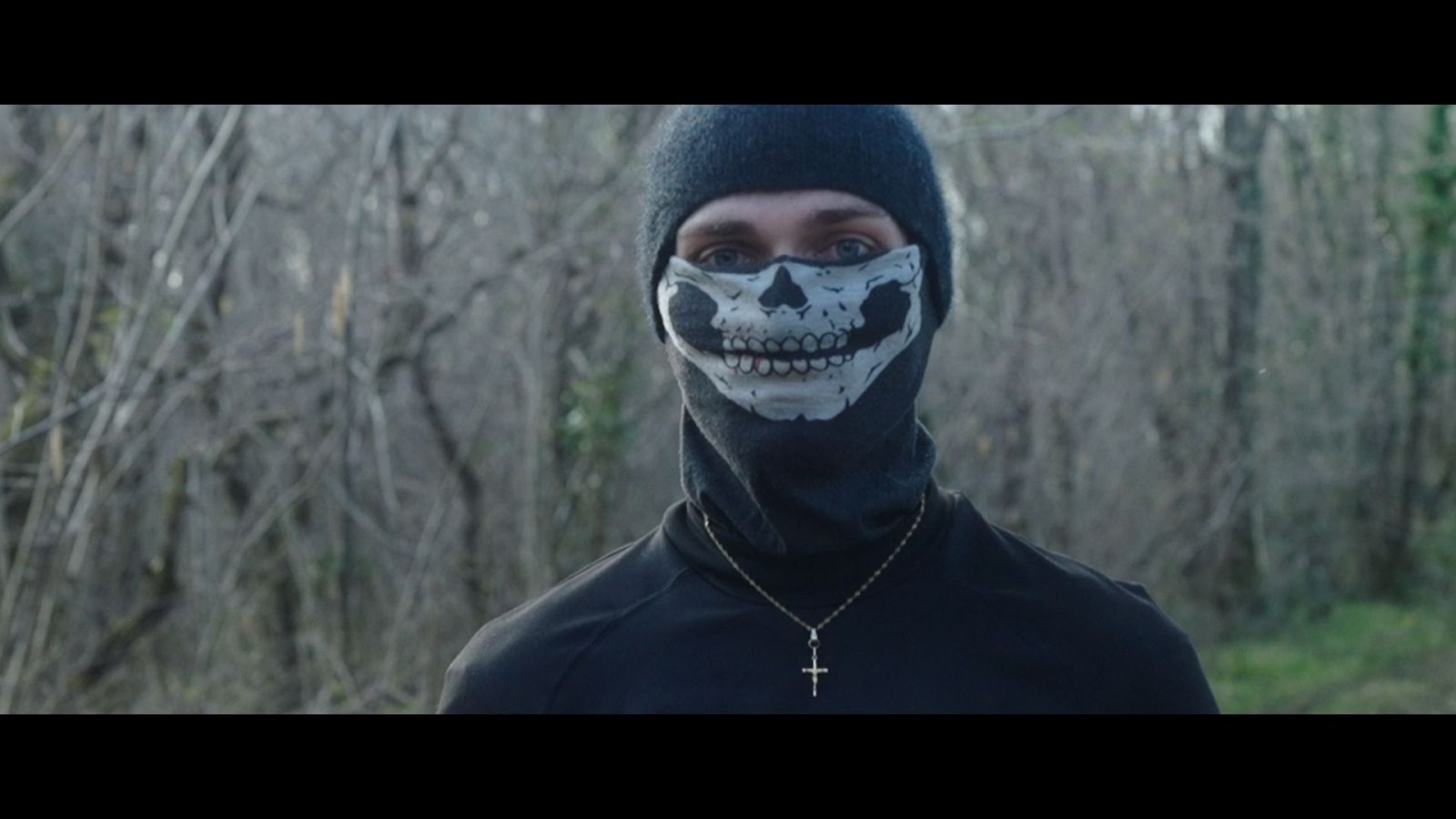 Un individuo con una máscara de esqueleto y vestimenta oscura posa al aire libre, con una cadena dorada y una cruz. El fondo muestra un paisaje natural con árboles y vegetación, sugiriendo una iluminación tenue.