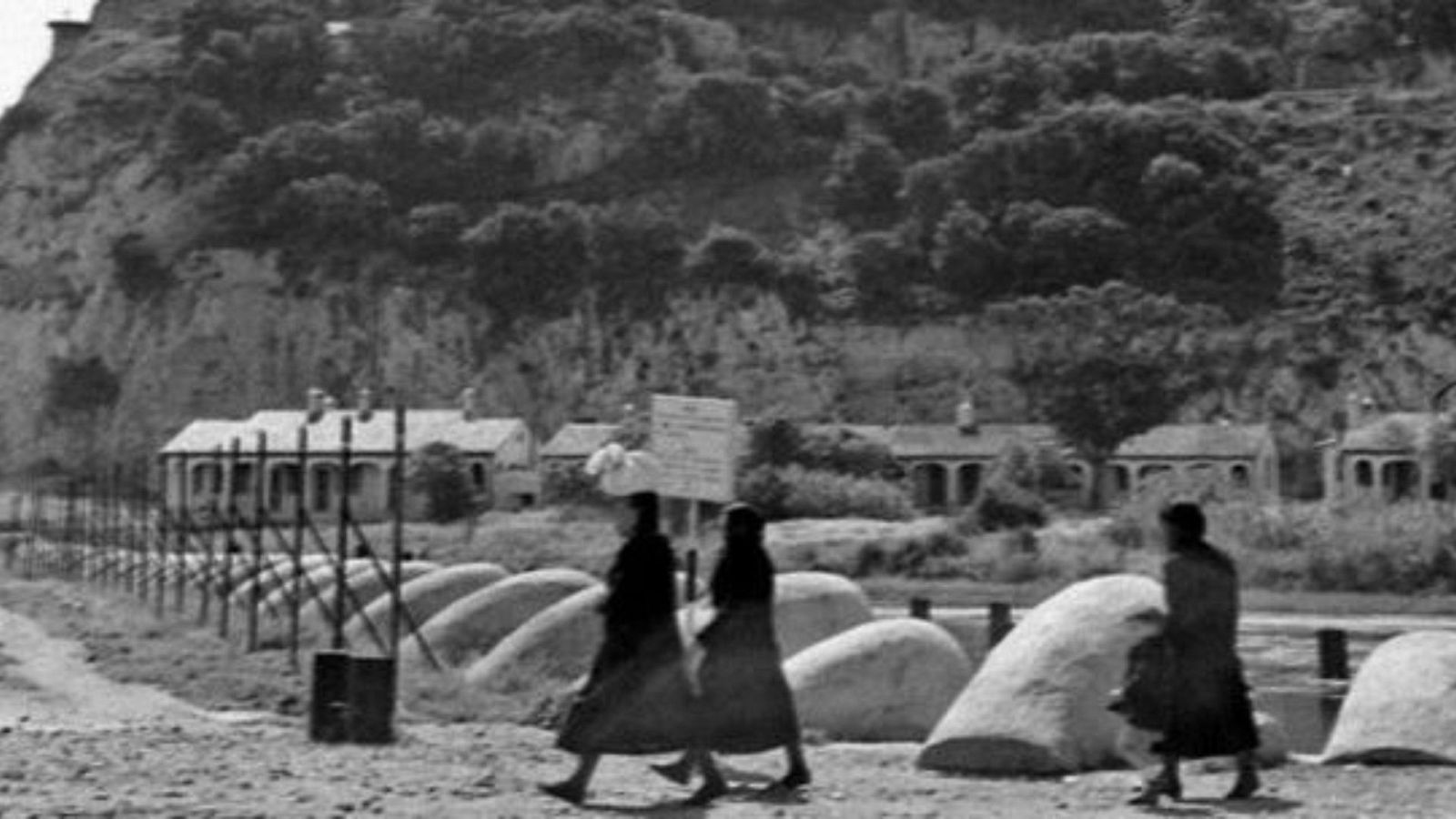 Una imagen antigua muestra a tres personas caminando por un camino en un entorno rural, con una colina y construcciones al fondo. Se observan rocas, una valla y un letrero con texto ilegible.