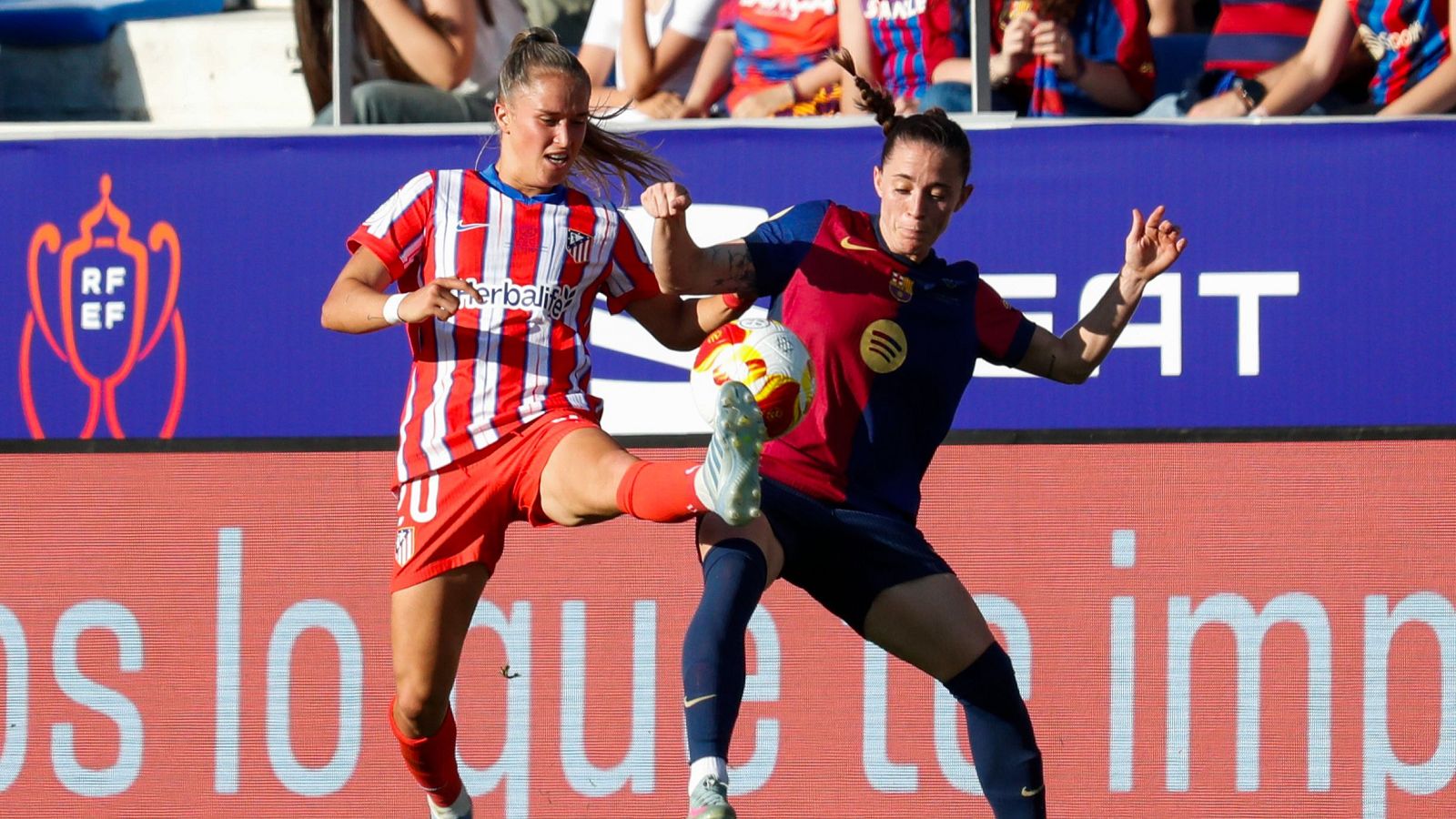 Disputa aérea por el balón entre dos jugadoras de fútbol femenino; una con camiseta rojiblanca y otra con azulgrana, mostrando concentración y esfuerzo.