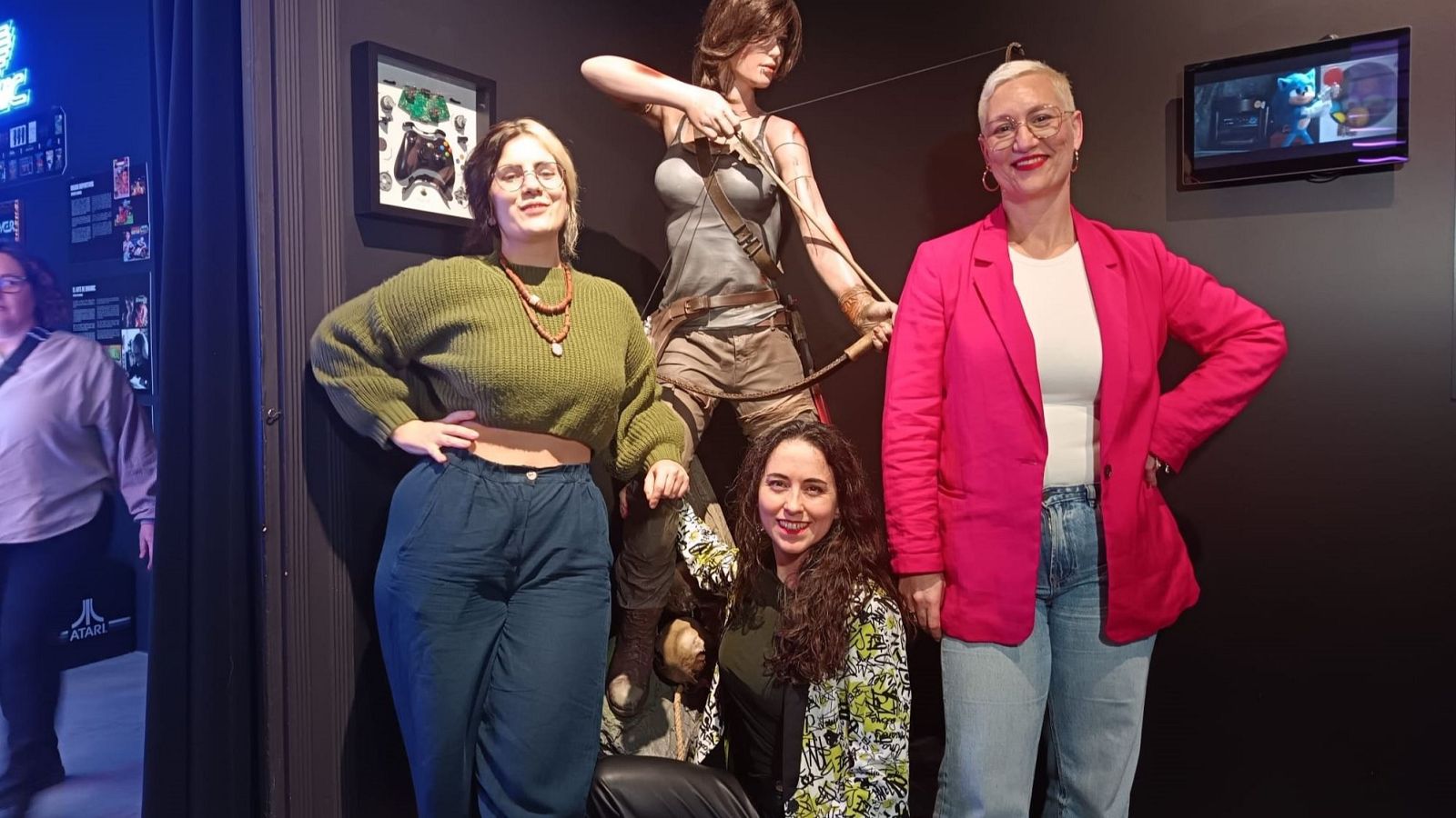 Tres mujeres se fotografían junto a una estatua de un personaje de videojuego, una de ellas con chaqueta rosa y gafas, otra con chaqueta estampada y la tercera con suéter verde.