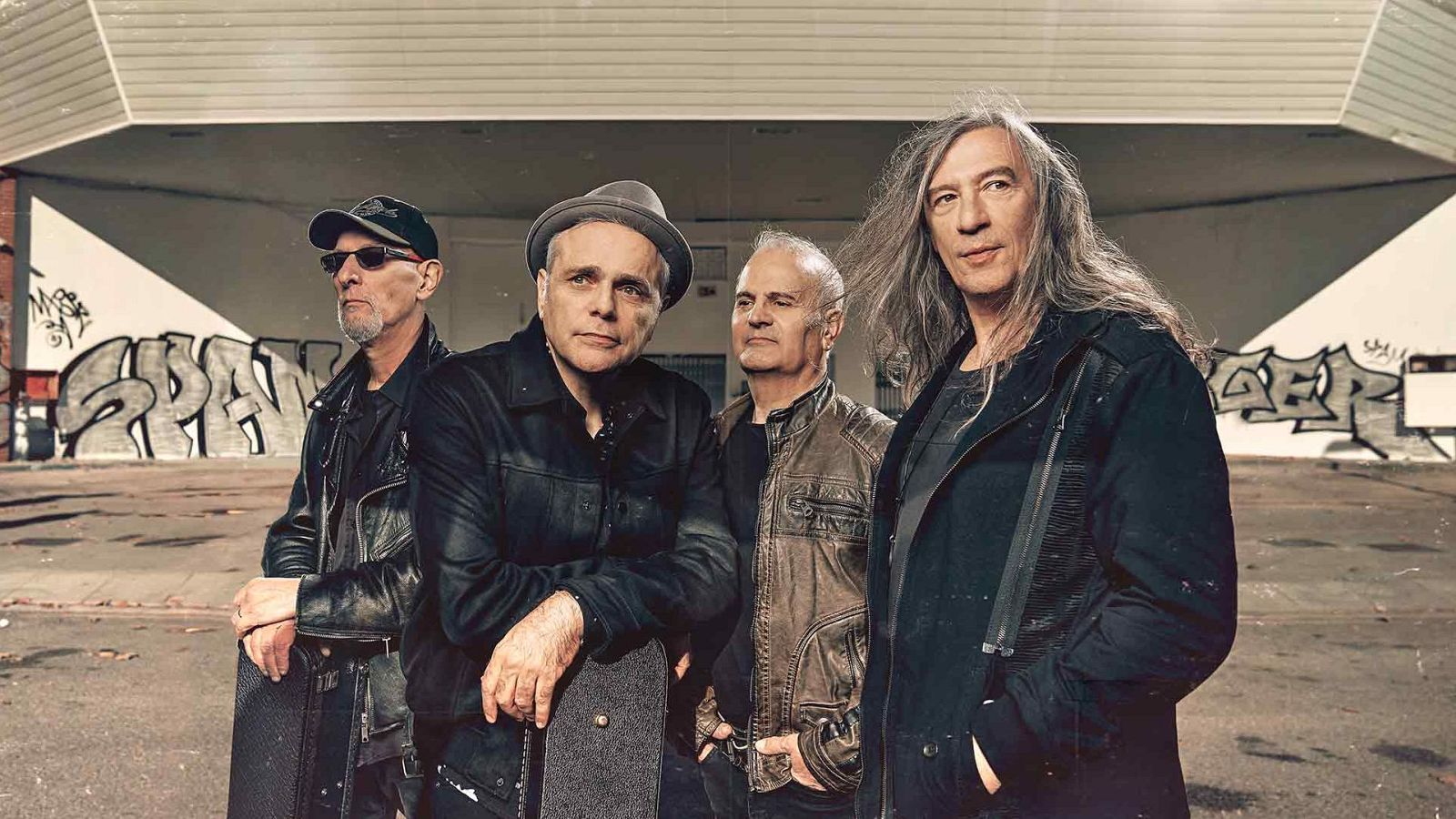 Quatre homes, presumiblement membres d'una banda musical, posen en un carrer amb grafitis. Vesteixen jaquetes de cuir i mostren un estil rocker, amb diferents complements com gorres, barrets i ulleres de sol.