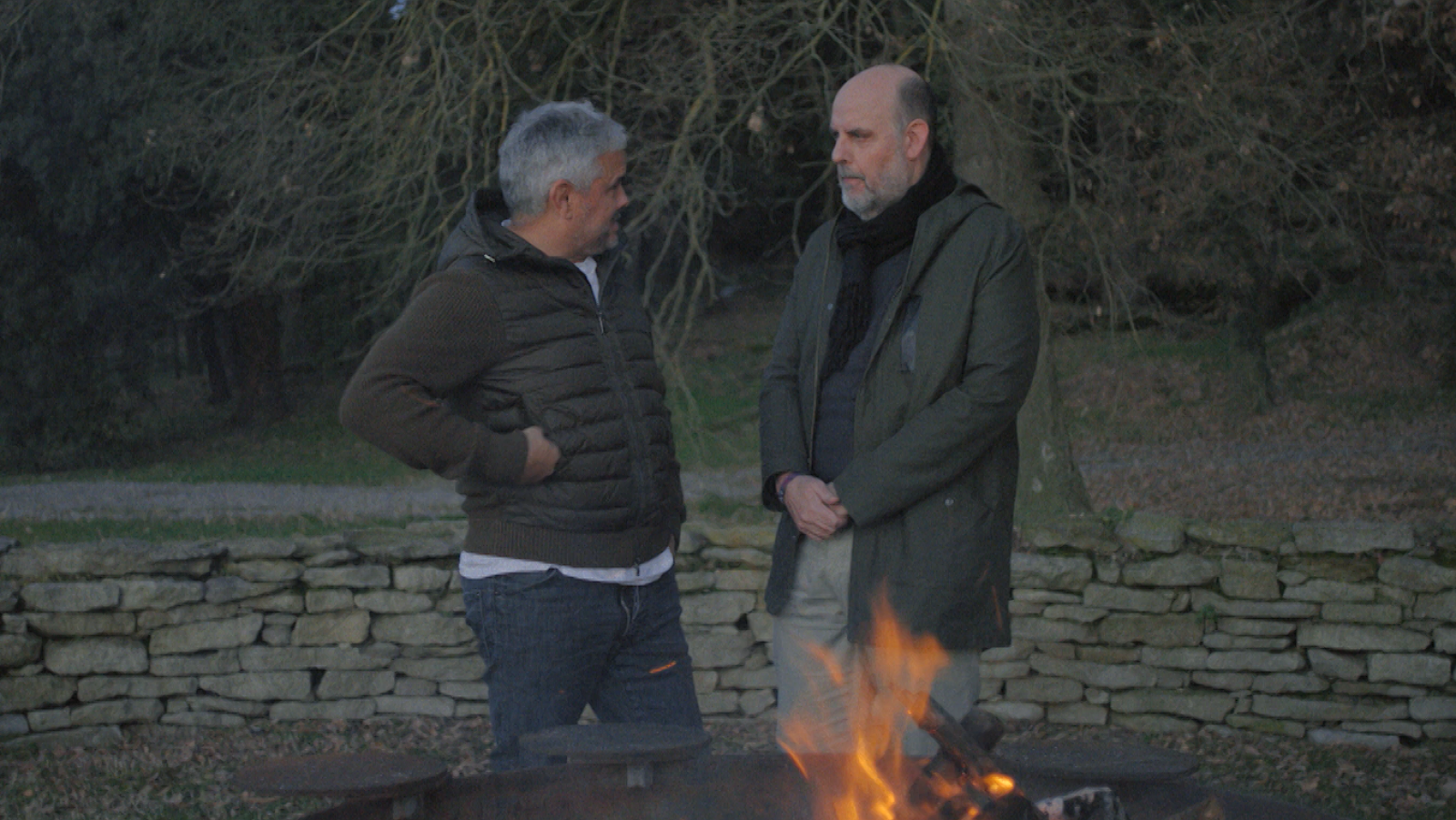 Dues persones conversen prop d'una foguera, un d'ells amb jaqueta fosca i l'altre amb abric llarg. Al fons, un mur de pedra i arbres sense fulles.