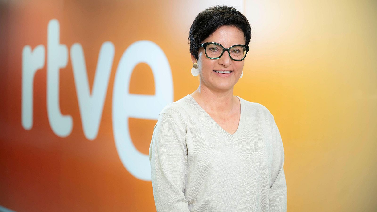 Marta Ribas, con gafas y un jersey claro, sonríe a la cámara. Detrás de ella, se aprecia un fondo difuminado con el logo de una cadena de televisión.