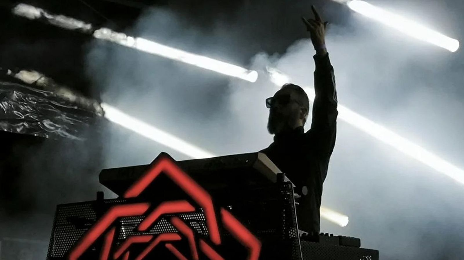 En un escenario iluminado, un artista con gafas de sol y barba, vestido de negro, realiza un gesto con la mano frente a su equipo musical, que presenta un diseño geométrico iluminado.