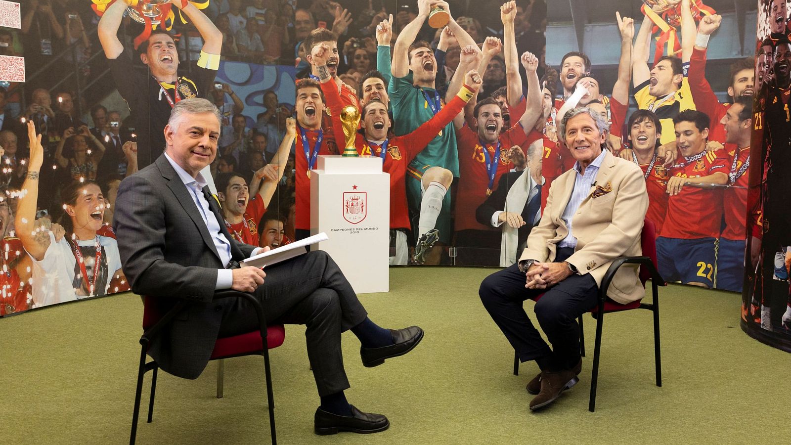 Dos hombres, uno con traje oscuro y otro con chaqueta clara, están sentados en un estudio frente a un fondo con imágenes deportivas. Un atril con un escudo se encuentra en primer plano.
