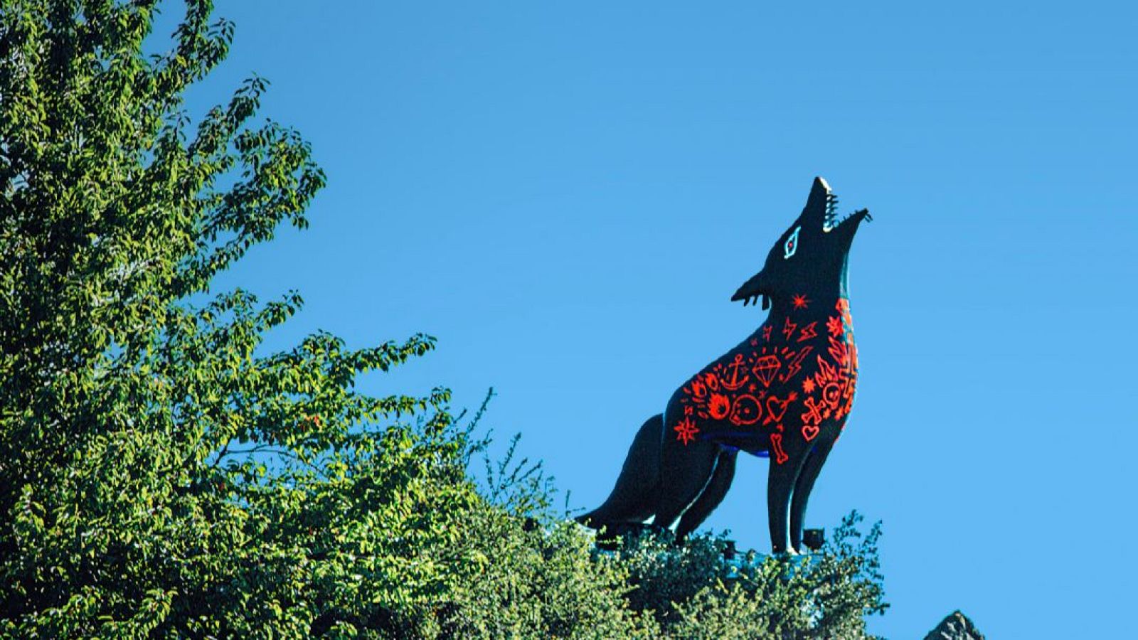 Una escultura de un lobo negro, adornada con símbolos rojos, se alza sobre sus patas traseras con la cabeza levantada. El fondo es un cielo despejado y un grupo de árboles.