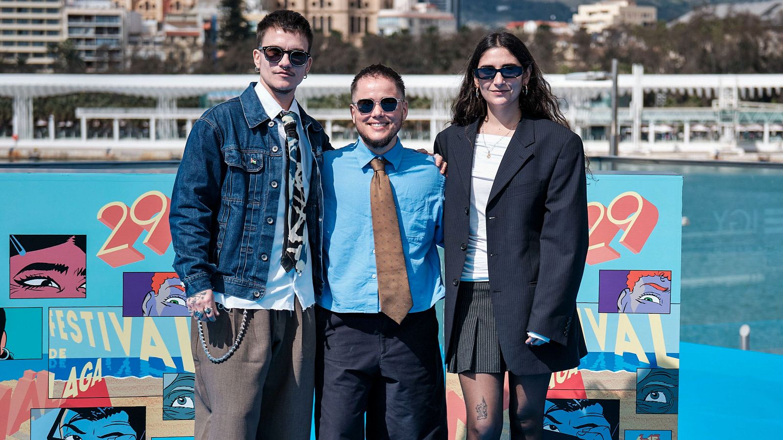 Un grupo de personas se fotografía delante de un cartel publicitario con el número '29' y la frase 'FESTIVAL DE MÁLAGA', con ilustraciones de ojos. Se distinguen detalles de la vestimenta de los individuos, como gafas de sol, chaquetas, camisas y corbatas.
