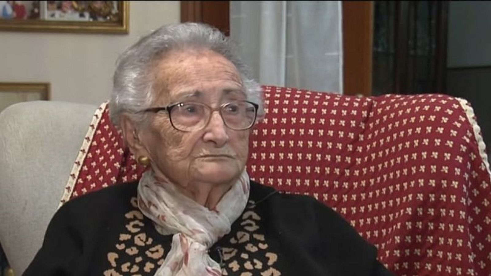 Isabel Oses, con gafas y bufanda floral, posa sentada en una silla con respaldo rojo. Se aprecia un jersey negro con estampado y pendientes dorados, en un ambiente interior con un cuadro en la pared.
