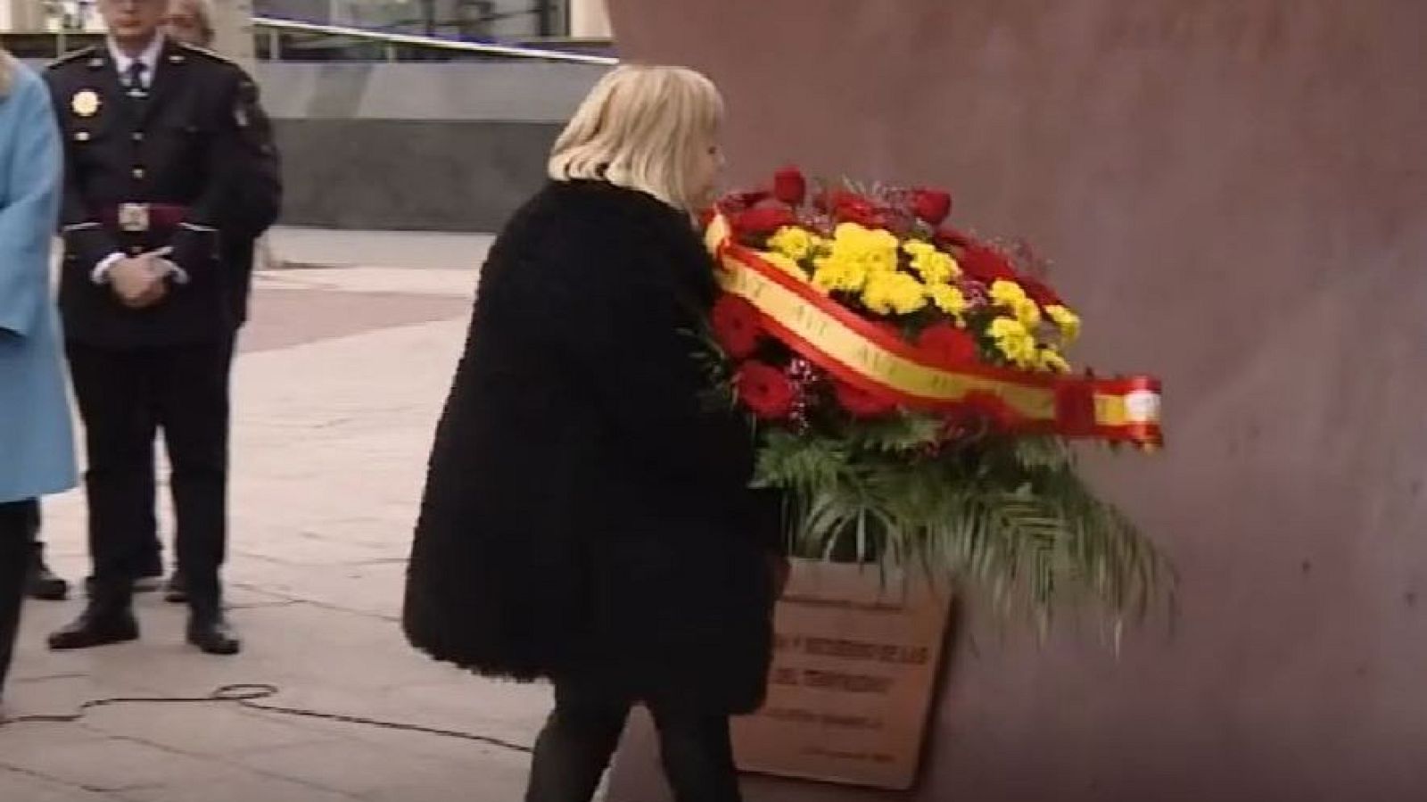 Una mujer rubia, vestida con un abrigo oscuro, coloca una corona floral con los colores de la bandera española. Un hombre uniformado de policía está presente en la escena, que tiene lugar en un entorno exterior.