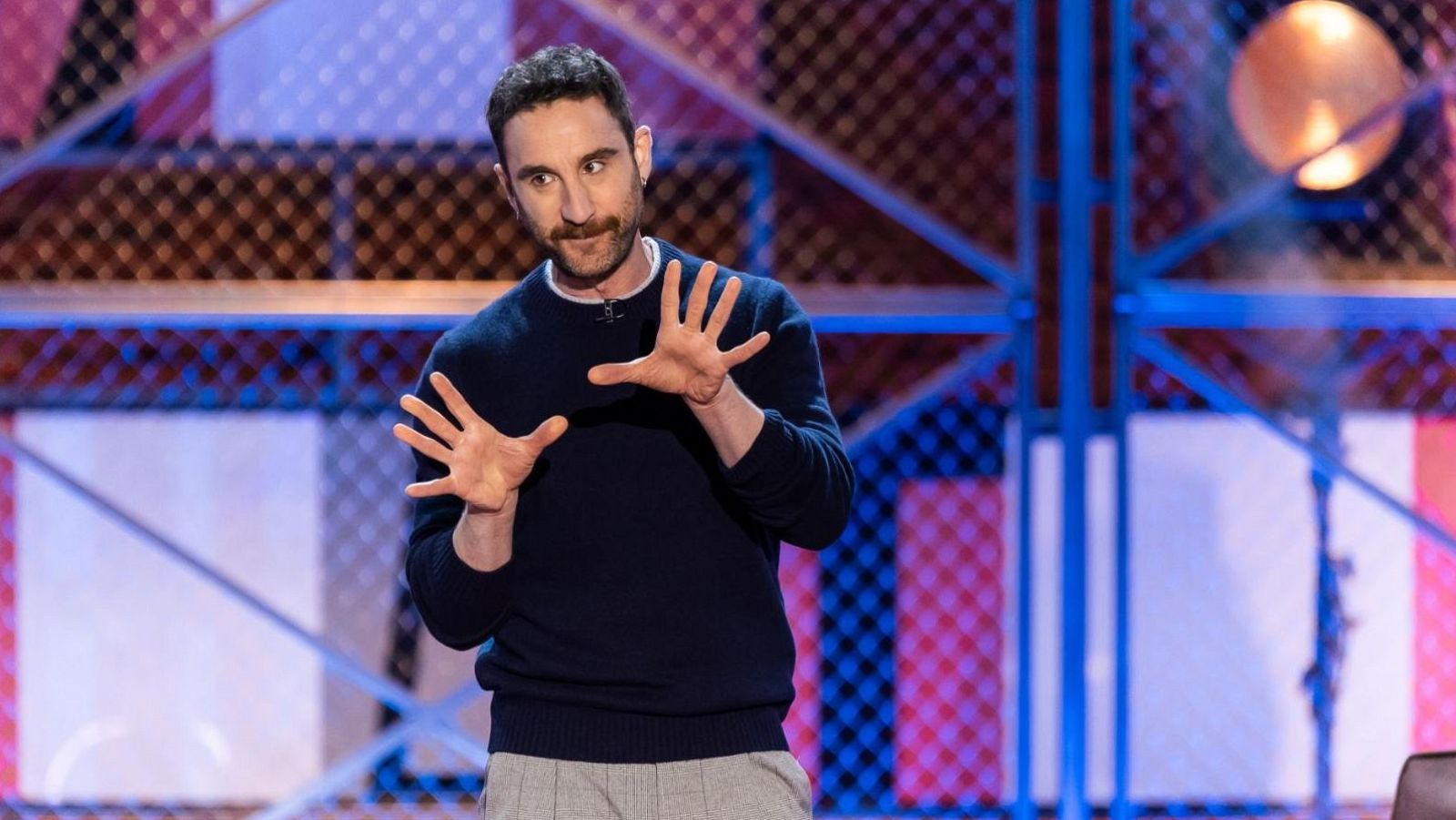 Dani Rovira, con jersey azul marino y pantalones grises, posa con las manos levantadas frente a un decorado con formas geométricas y una estructura metálica azul.