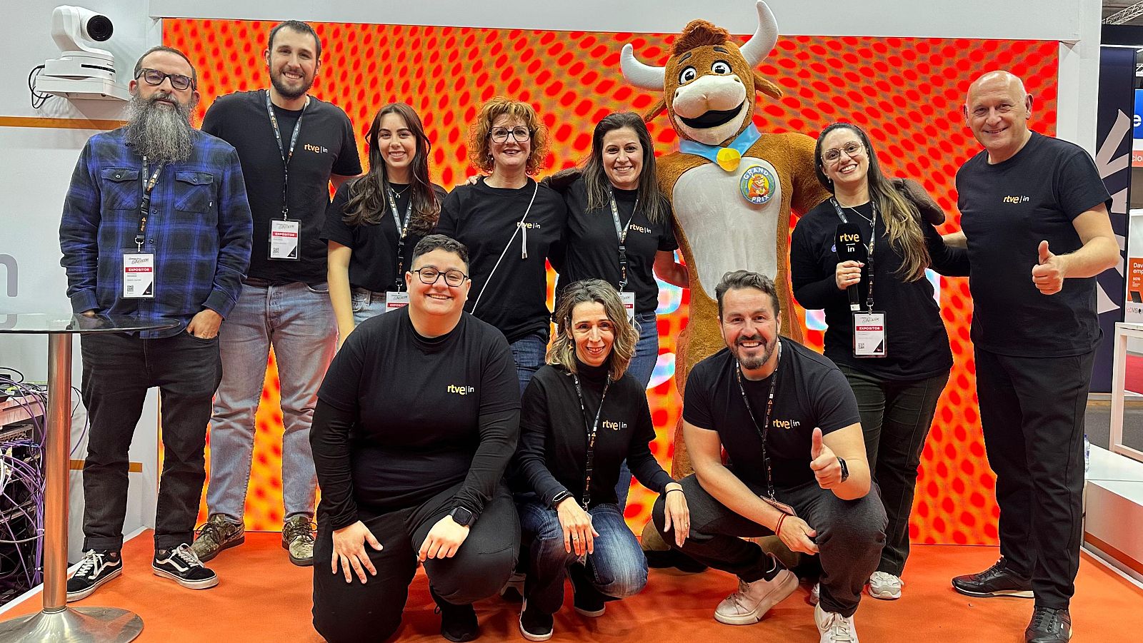 Un grupo diverso de personas, incluyendo a alguien disfrazado de toro, posa para una foto. Todos visten camisetas negras con un logo, frente a un fondo abstracto en tonos cálidos.
