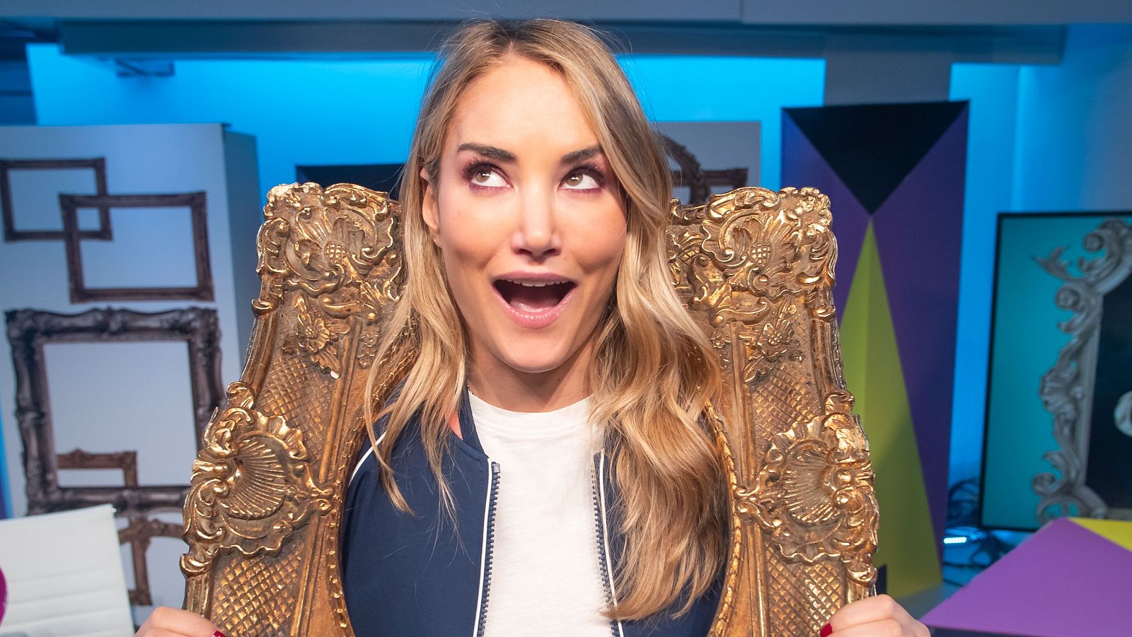 Una mujer rubia, con una expresión de sorpresa, sostiene un marco dorado ornamentado. El fondo muestra otros marcos y formas geométricas.