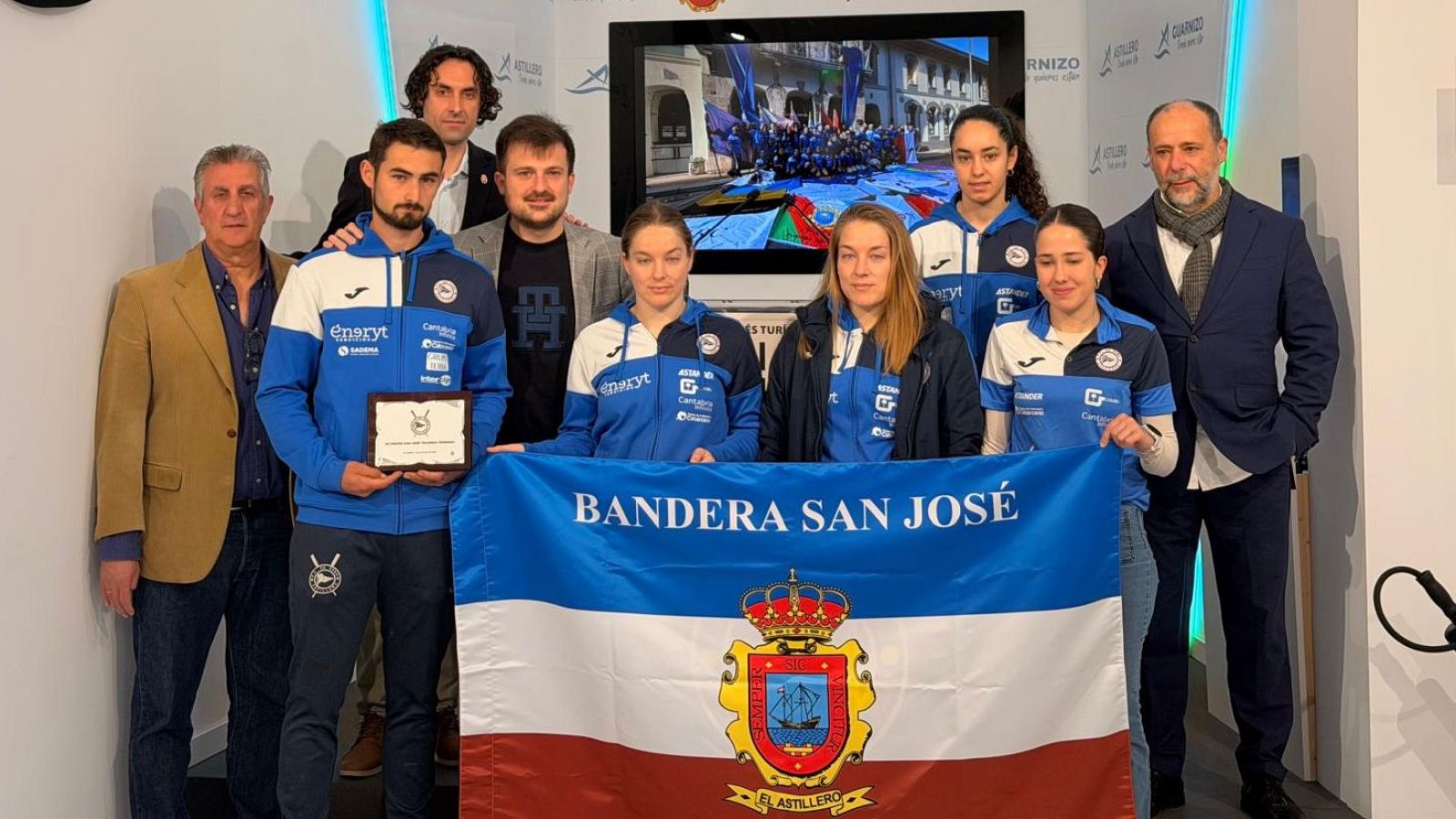En una sala, un equipo femenino de Astillero celebra con un trofeo y una bandera. Las deportistas visten remeras con el logo de su equipo y patrocinadores, mientras que detrás de ellas se encuentran otras personas, incluyendo a un hombre con traje.