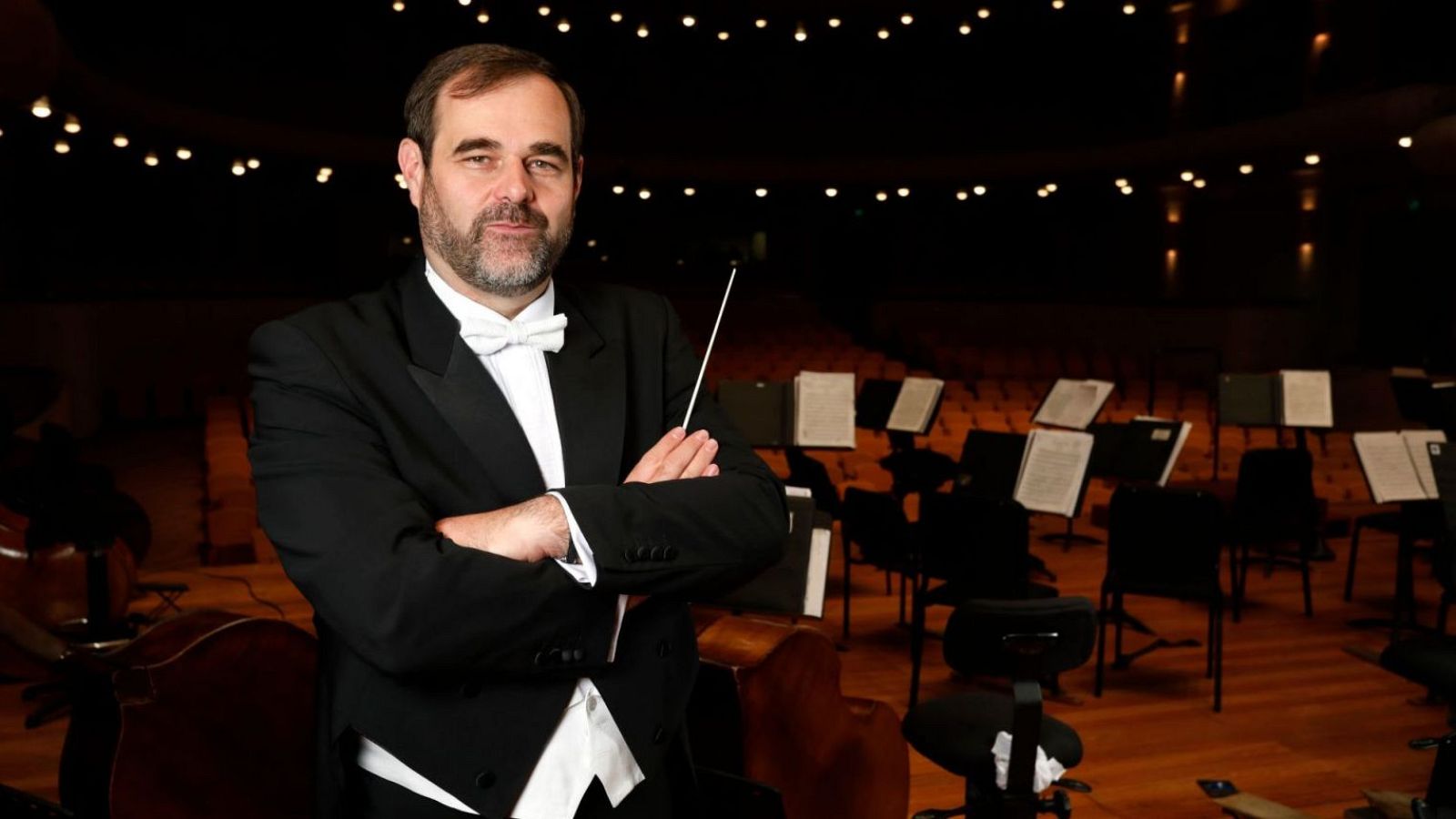Josep Caballé, vestido con un esmoquin negro y pajarita blanca, posa con una batuta en un escenario iluminado. Se observan atriles con partituras y algunos instrumentos musicales.