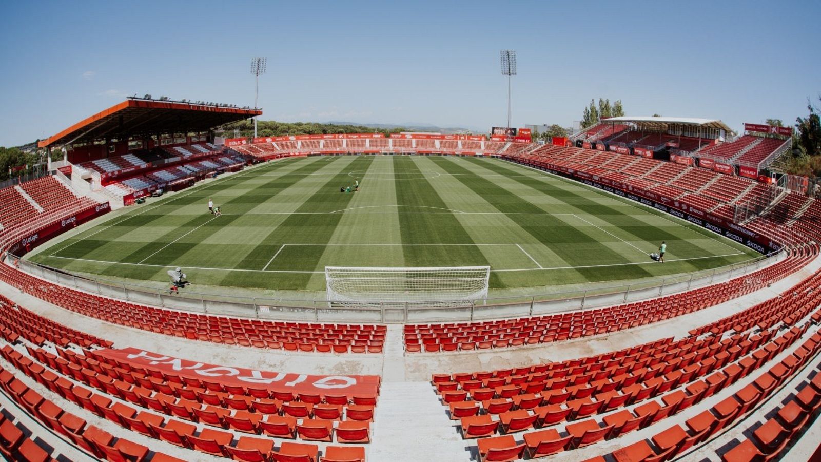 L'estadi presenta grades vermelles i un camp de gespa amb ratlles, amb porteries a banda i banda. Hi ha personal treballant al camp, mentre que les grades estan majoritàriament buides.