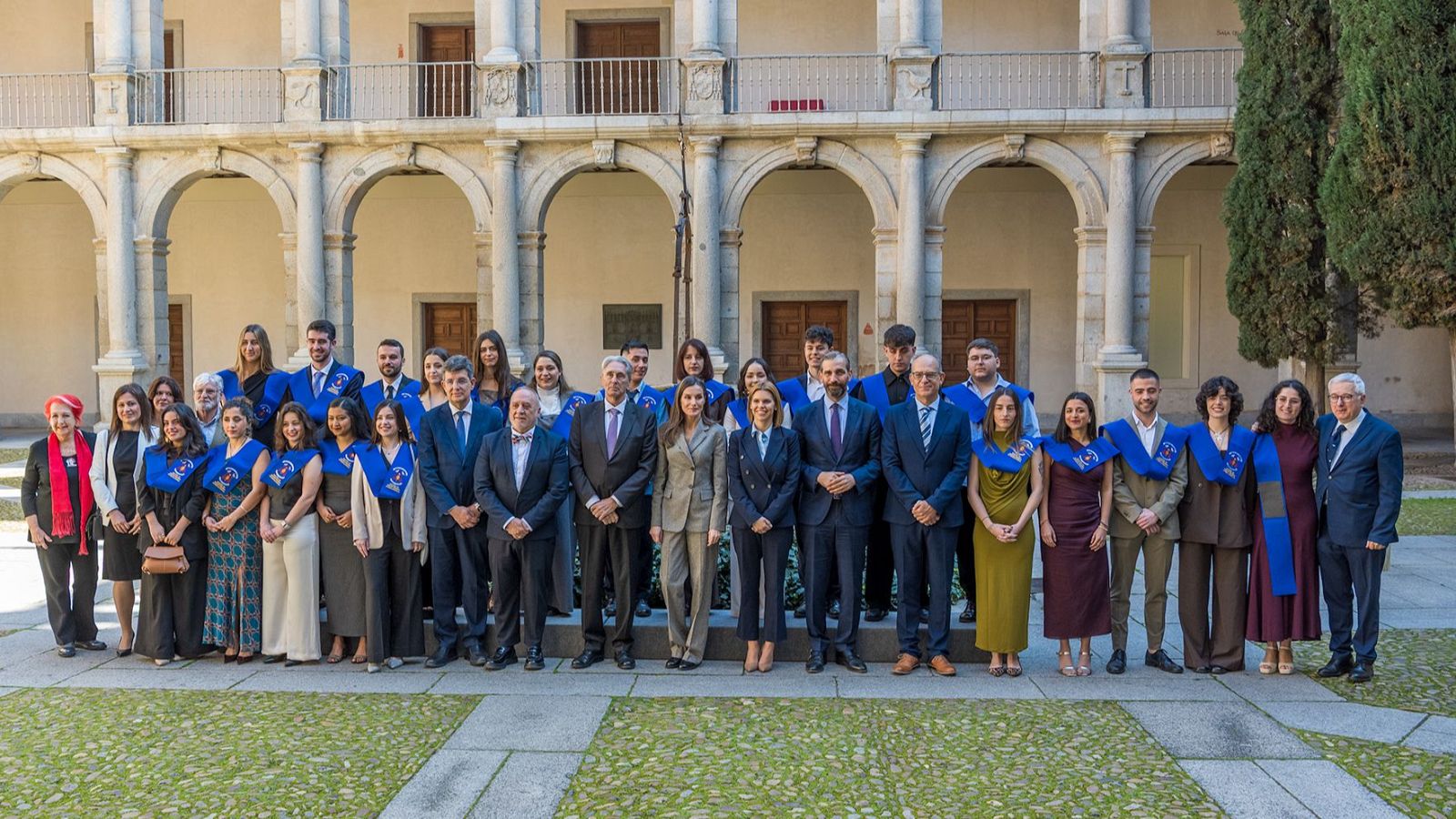 Un grupo de personas posa en un patio con arcos de piedra. En el centro, una mujer con traje gris está rodeada de personas vestidas formalmente, algunas con estolas azules.