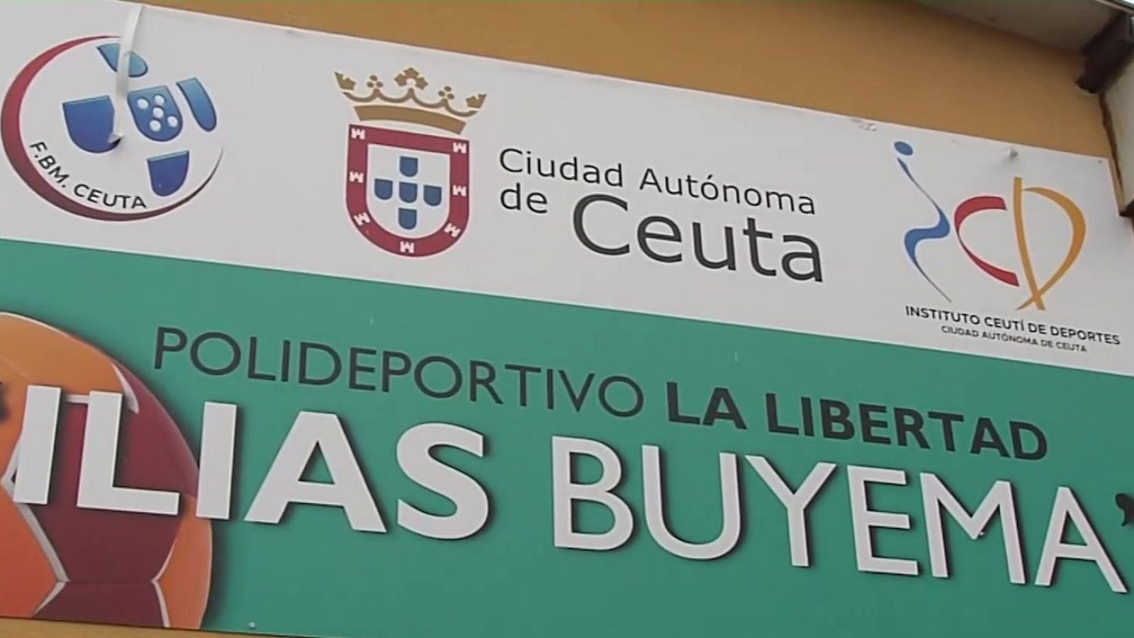 Un letrero informativo en Ceuta anuncia el Polideportivo La Libertad Ilias Buyema, con los logos de varias instituciones en la parte superior y el nombre del polideportivo en letras grandes sobre un fondo verde, incluyendo una imagen de un balón de fútbol.