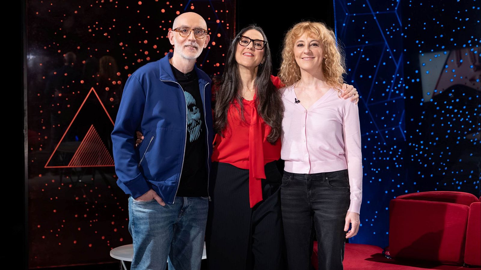 Tres personas posan para una foto: un hombre con gafas y camiseta estampada, una mujer con blusa roja y falda negra, y otra mujer con chaqueta rosa y vaqueros. El fondo presenta formas geométricas y sillones rojos.