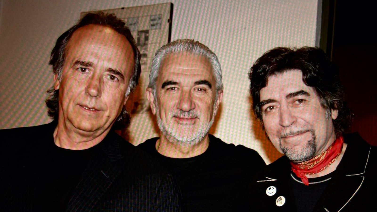 Tres hombres posan para la foto. Uno de ellos lleva una chaqueta negra y tiene el pelo castaño oscuro. El hombre del centro, con pelo y barba canosos, viste una camiseta negra. El tercero, con pelo rizado oscuro, lleva una chaqueta negra con pines y un pañuelo rojo.
