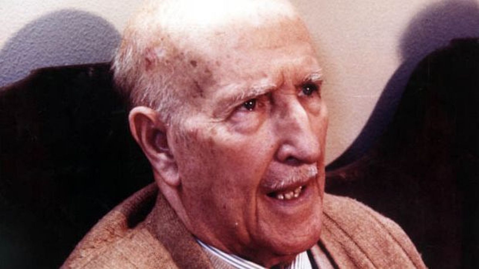 En primer plano, se aprecia el rostro de un hombre mayor con calvicie, cejas pobladas y bigote blanco. Viste una camisa a rayas y una chaqueta clara, con una pared blanca como fondo.