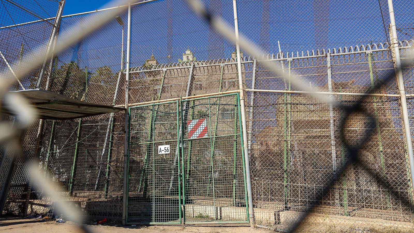 Un paso fronterizo cerrado se muestra con múltiples vallas metálicas y alambre de púas. Una puerta verde con señalización de peligro impide el acceso, mientras que un edificio clásico se vislumbra al fondo, en un día soleado.