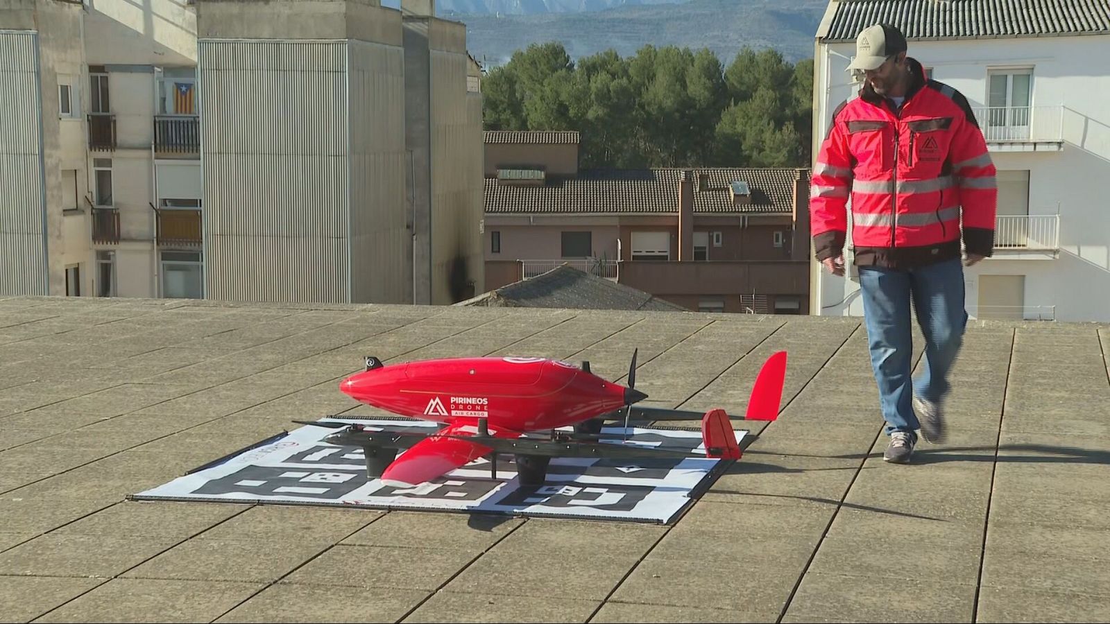 Un dron rojo de la marca "PIRINEOS DRONE" se sitúa sobre una plataforma. Un hombre con ropa de alta visibilidad camina cerca, con edificios y árboles de fondo.