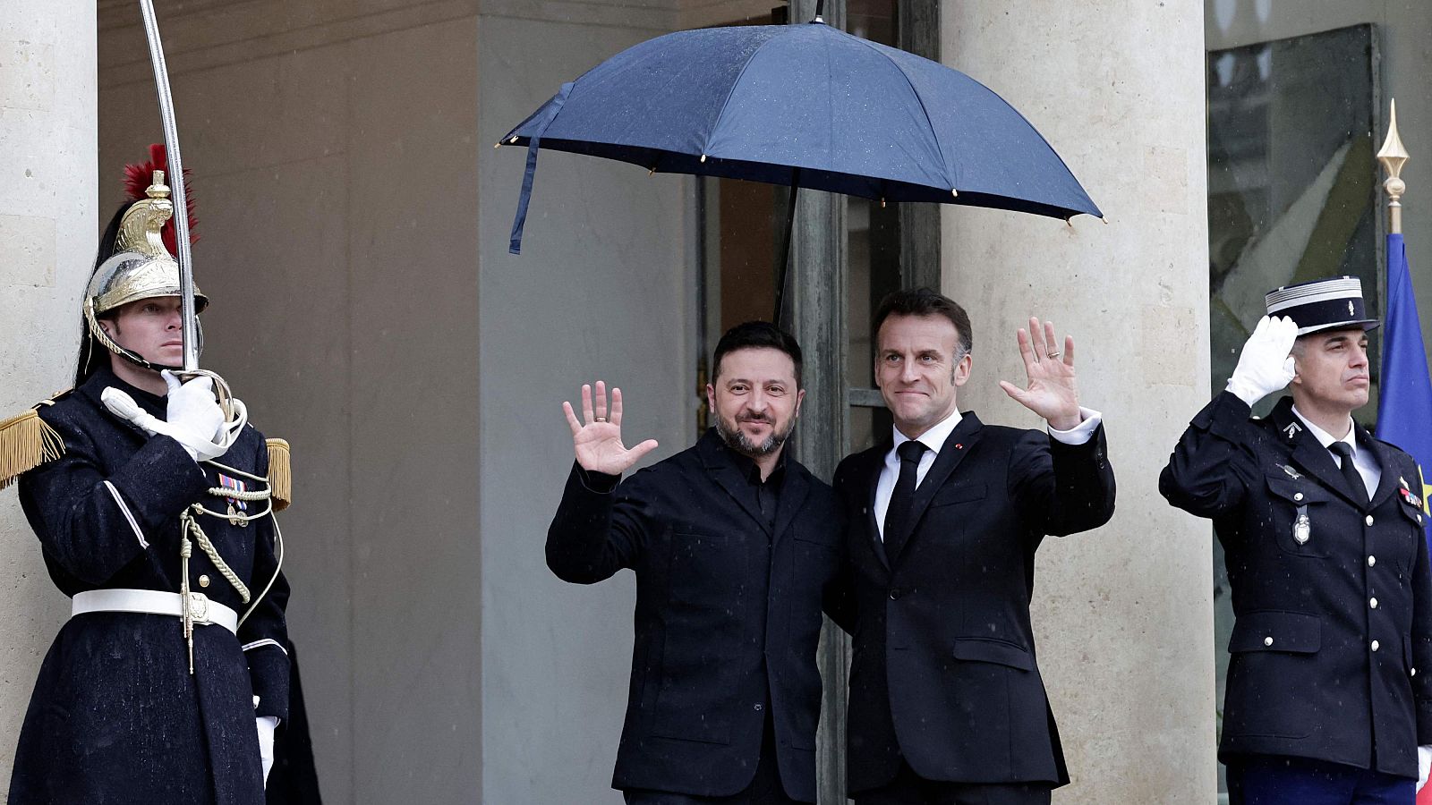 Emmanuel Macron y Volodímir Zelenski se encuentran en el exterior de un edificio, sonriendo y saludando. Dos guardias uniformados, uno de ellos con espada, flanquean a los mandatarios bajo un paraguas.