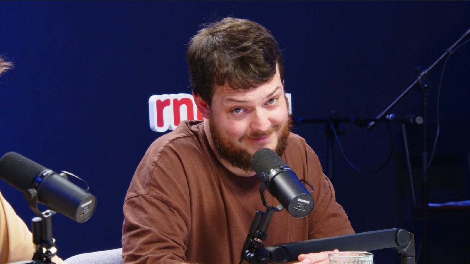 En un estudio con fondo azul oscuro, Xavi Daura, con barba y camiseta marrón, se encuentra frente a un micrófono negro, posiblemente grabando un podcast.