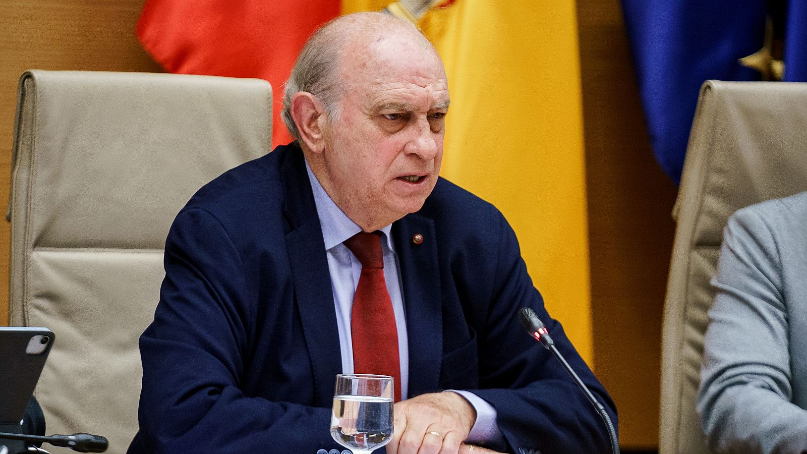 Jorge Fernández Díaz, en traje azul marino y corbata roja, durante una comparecencia.  Habla ante un micrófono, con un vaso de agua cerca.