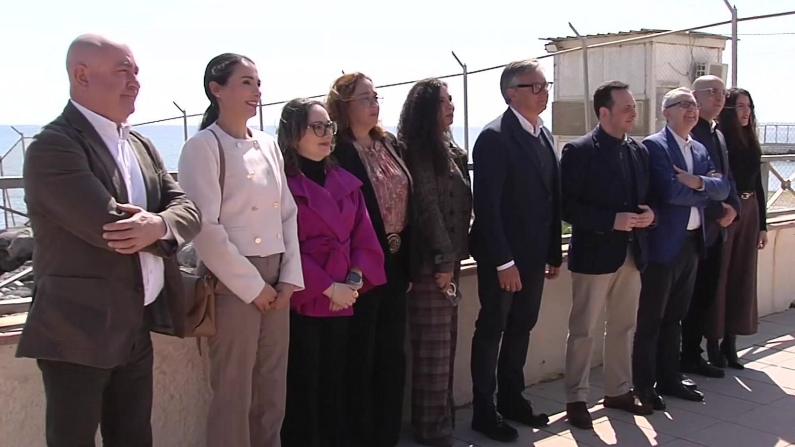 Un grupo diverso de personas, incluyendo hombres con trajes y mujeres con diferentes estilos de ropa, posan para una foto al aire libre, con una valla y vistas al mar en el fondo.