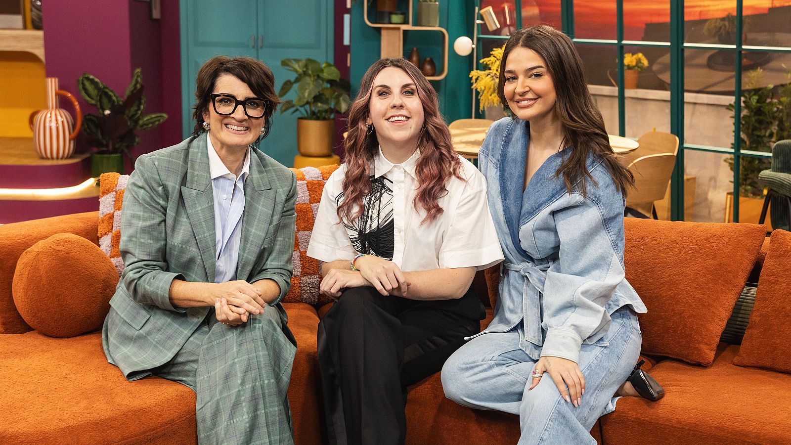 Tres dones, incloent una amb vestit a quadres i una altra amb camisa blanca, estan assegudes en un sofà taronja en un estudi de televisió amb decoració de fusta i plantes.