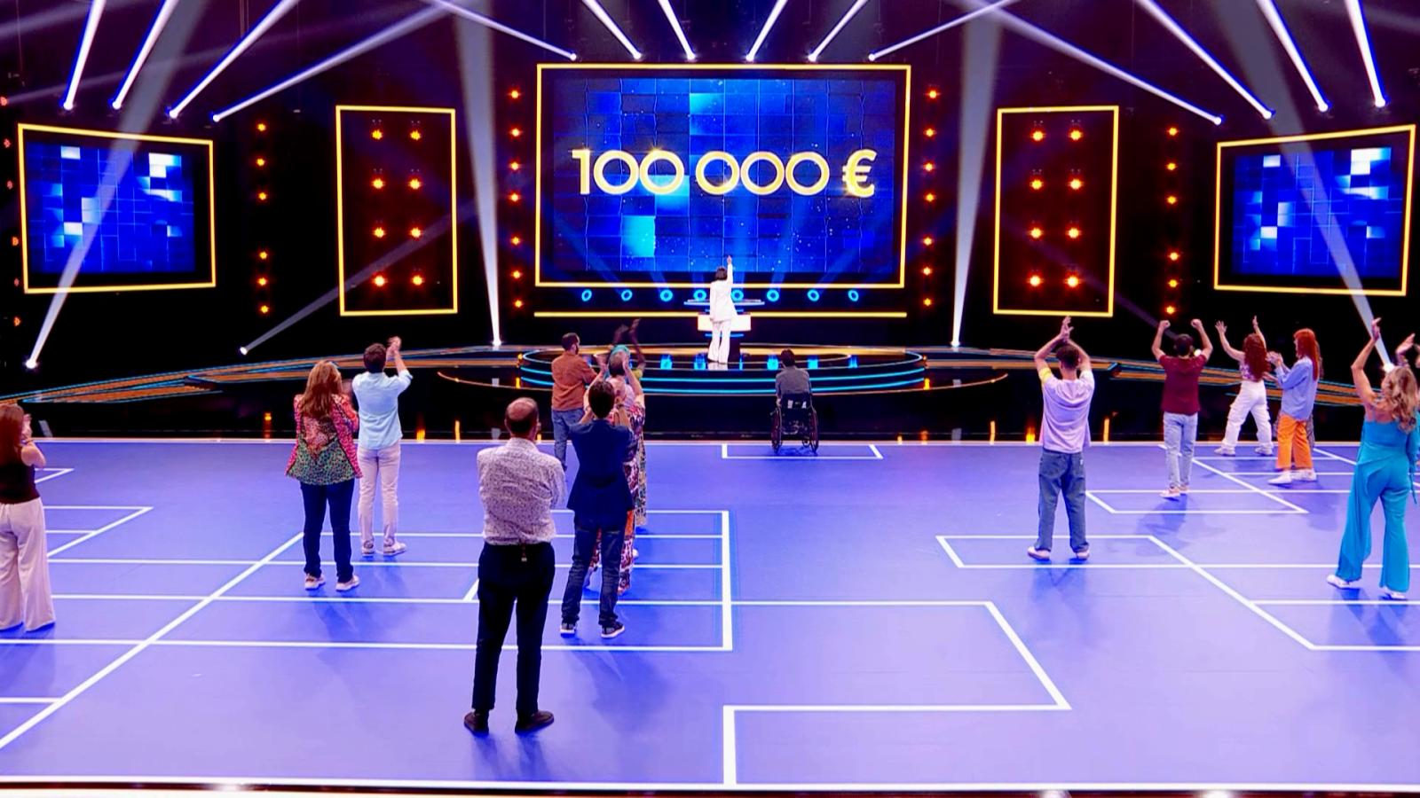 En el estudio de televisión, el presentador, vestido de blanco, se dirige a los concursantes mientras una pantalla muestra el premio. Un grupo de concursantes celebra en el suelo, incluyendo a una persona en silla de ruedas, en un ambiente de emoción.