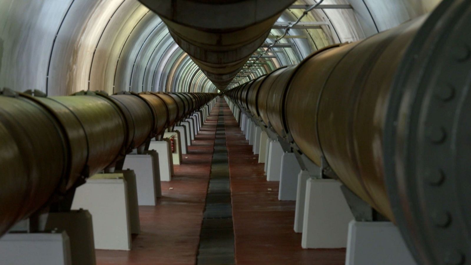 Vista interior d'una depuradora d'aigua, amb un túnel arquejat ple de canonades marrons i suports blancs, amb parets de formigó gris clar i un terra marró vermellós.