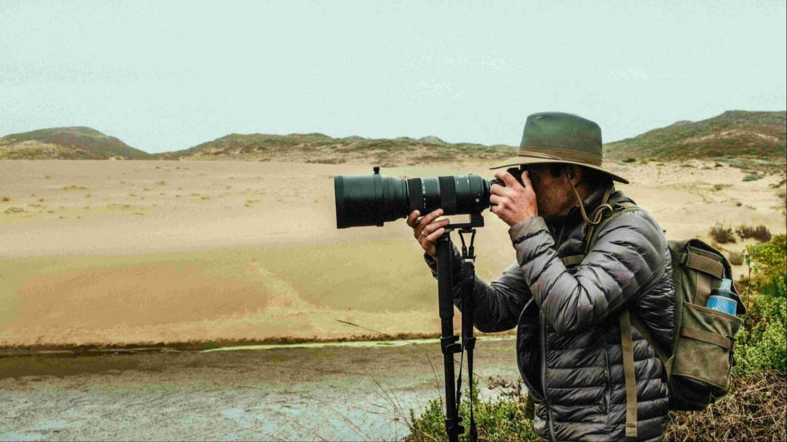 Un fotógrafo con sombrero y chaqueta gris, equipado con una cámara profesional y teleobjetivo sobre un trípode, se prepara para fotografiar un paisaje costero con playa, agua y montañas, bajo un cielo nublado.