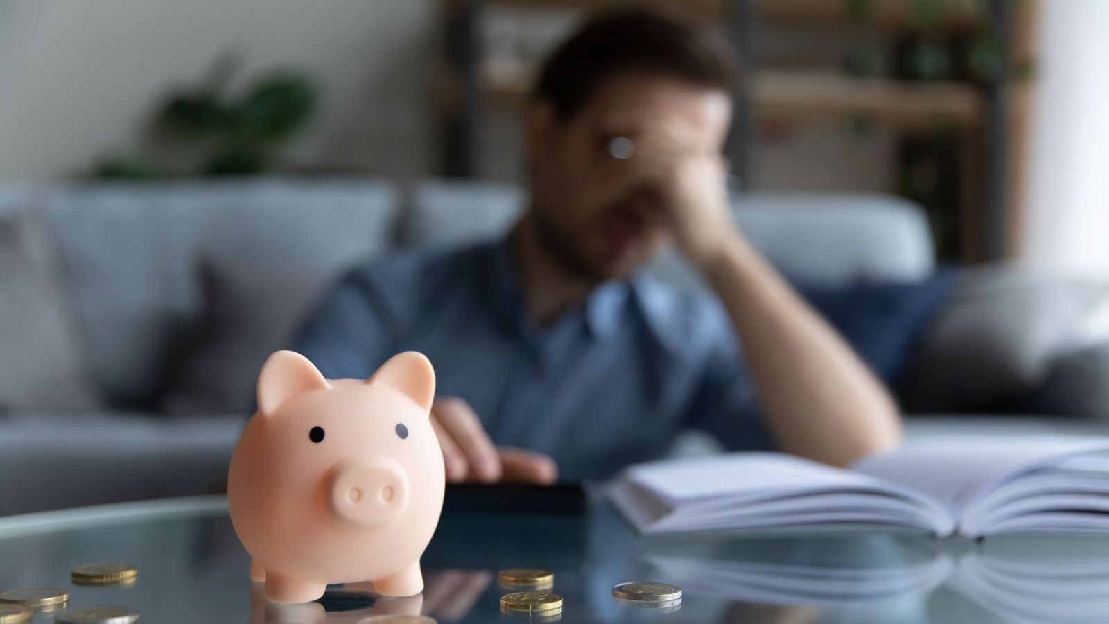 Una mesa en la que aparecen un hombre sentado con las manos tapándose la cara  junto a una libreta de fondo y una hucha de un cerdito con monedas alrededor