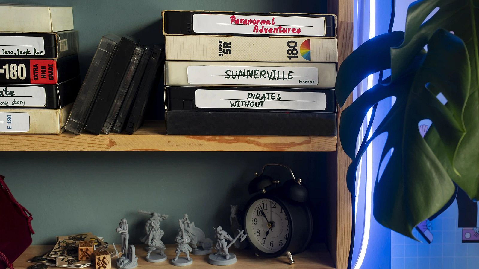 Una estantería de madera con dos niveles exhibe cintas VHS con títulos legibles y libros oscuros en la parte superior, mientras que la inferior muestra figuras en miniatura y un reloj despertador.