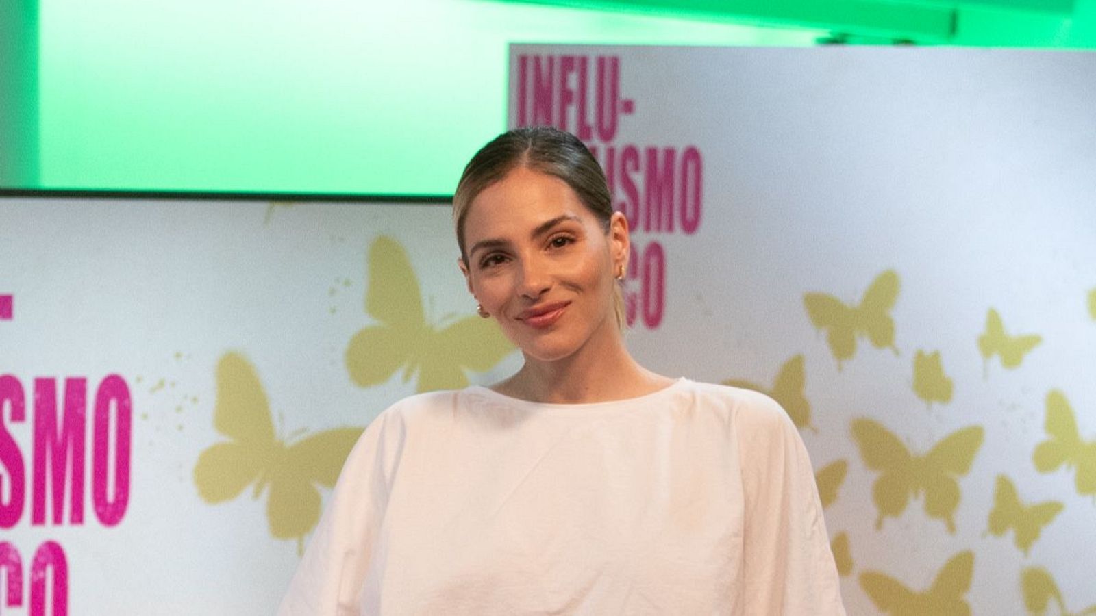 Andrea Duro, con el pelo recogido y una blusa blanca, posa frente a un fondo con mariposas amarillas y el título de un evento en letras rosas.