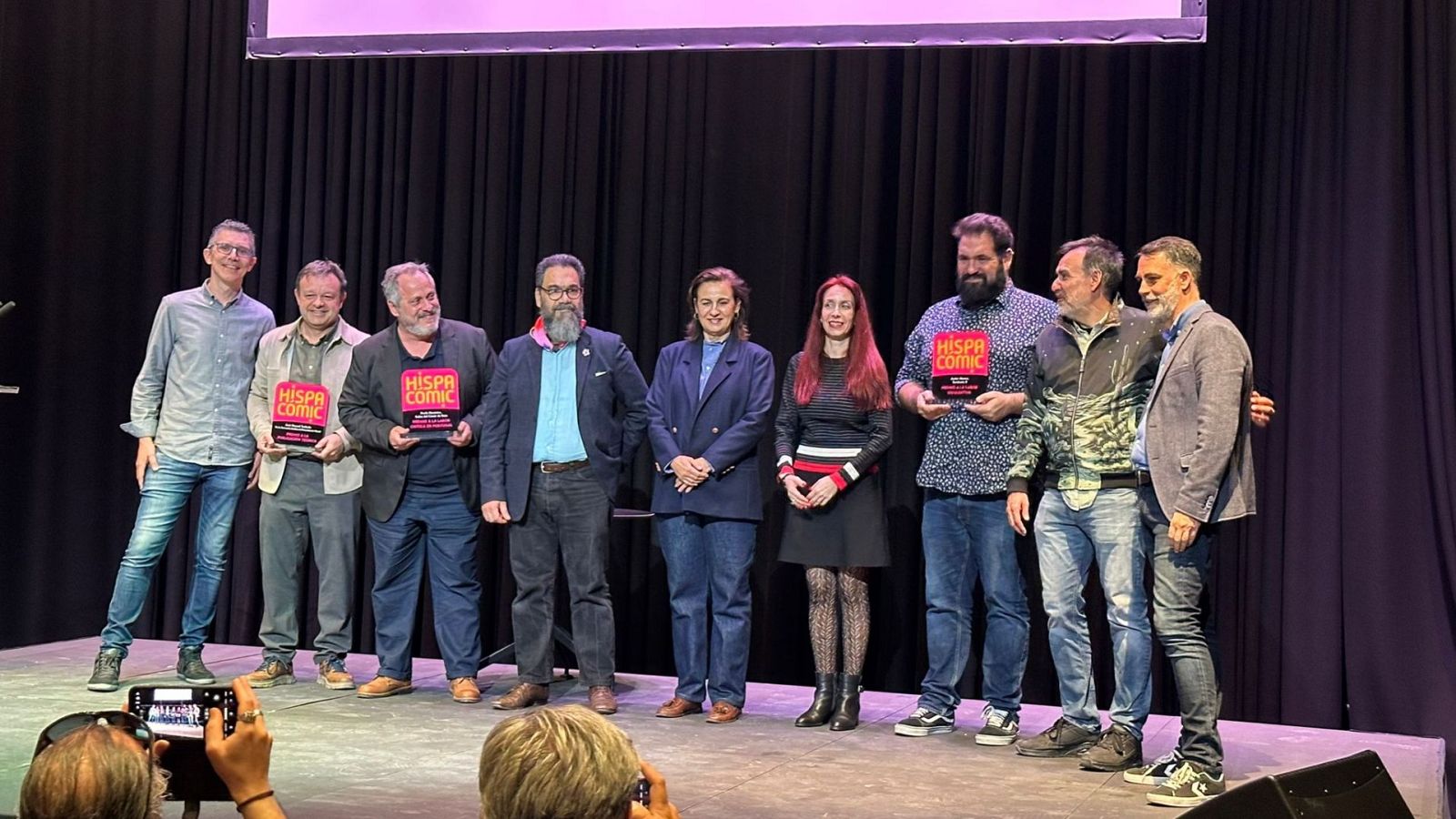 Un grupo de personas, posiblemente ganadores, posan en un escenario sosteniendo premios con el logo de "Hispacómic", mientras alguien toma una foto con su móvil.
