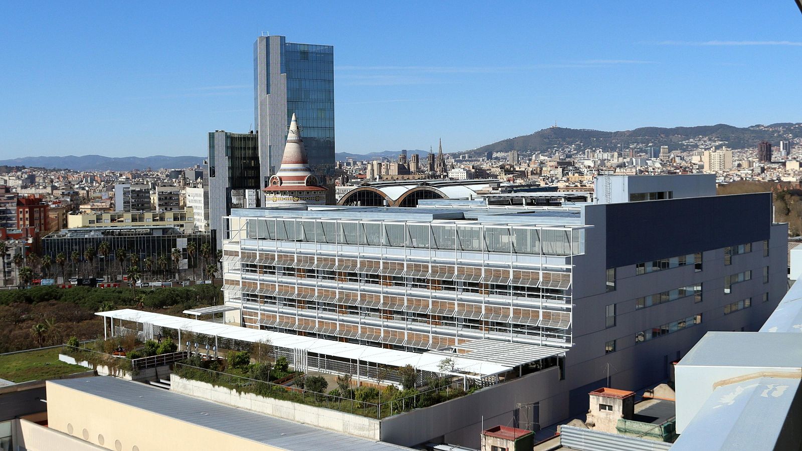 Una vista panorámica de Barcelona muestra el Hospital del Mar en primer plano, un edificio moderno con numerosas ventanas y una sección gris oscura, con el horizonte de la ciudad y la Sagrada Familia al fondo.